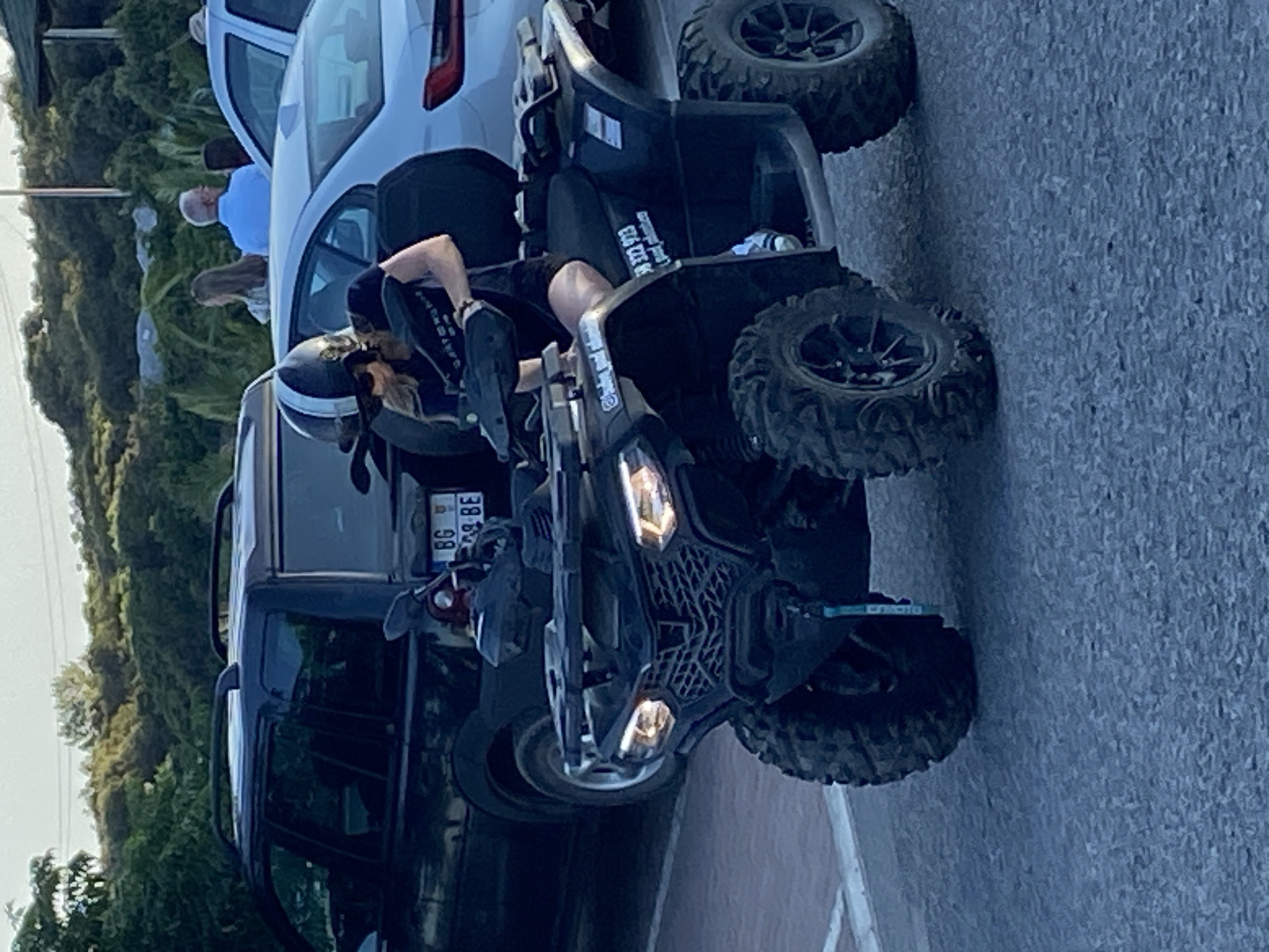 A woman wearing a black helmet and sunglasses is seated on a black ATV. She is wearing a black t-shirt and black shorts. The ATV has a license plate that reads "BG 98 BE". In the background, there are cars, including a black SUV and a silver sedan. People are visible in the distance. The scene appears to be a street in Prijevor, Montenegro.