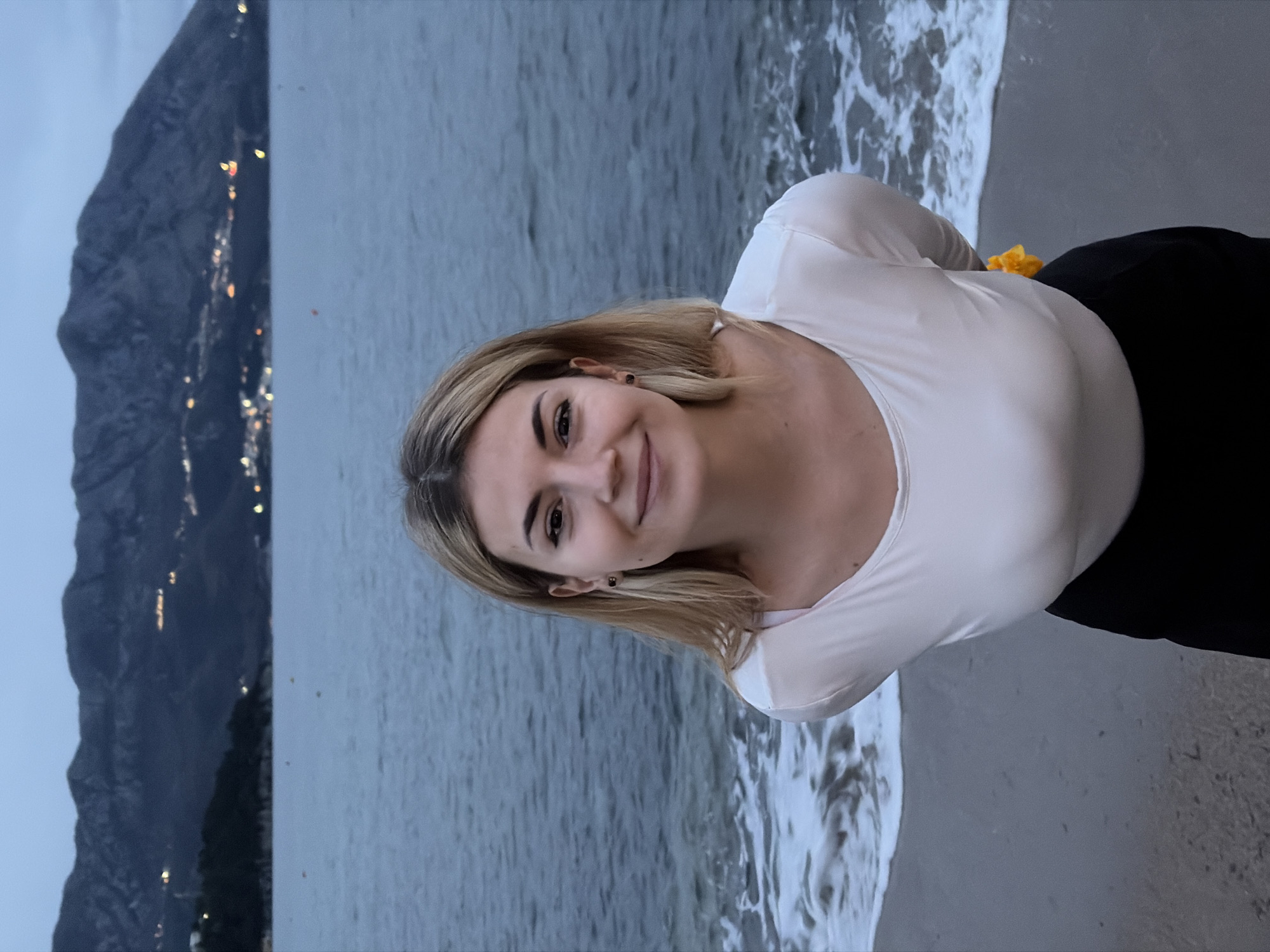 The image shows a young woman smiling at the camera. She has blonde hair and is wearing a white long-sleeved shirt and black pants. Behind her is the sea, with waves washing onto the sand. In the background, on the left, are mountains. Scattered lights on the mountainside suggest a city at night. Based on the mountainous backdrop and coastal environment, this is likely a beach in or near Budva, Montenegro.