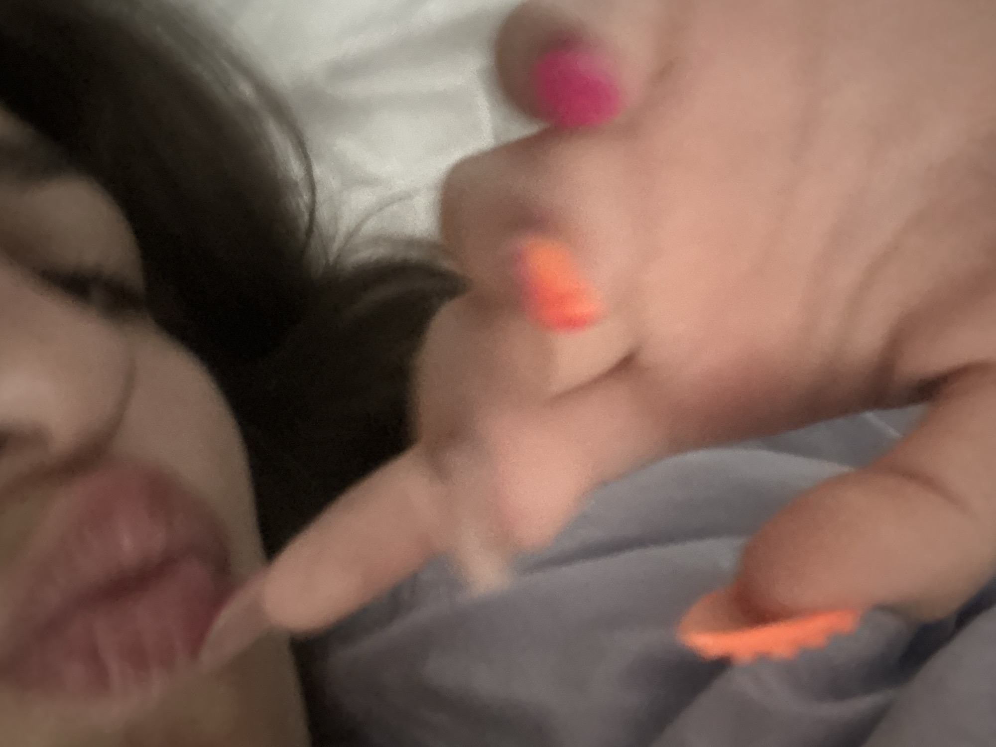 A close-up, low-angle shot captures a person's face and hand against a blurred background, possibly a bed. The person's left side of their face is visible, with dark hair framing their features. Their mouth is slightly open and appears to be the focal point, with full, reddish lips. A finger from their right hand, adorned with a bright pink nail polish, is extended towards the mouth, seemingly in the act of touching or pointing. Another finger on the same hand has vibrant orange nail polish, also visible in the frame. The skin appears soft and smooth. The overall impression is intimate and personal, with the background being indistinct, suggesting a private setting. The scene is devoid of any specific urban or natural landmarks that would place it in Stațiunea Zoologică Marină Agigea, Romania.