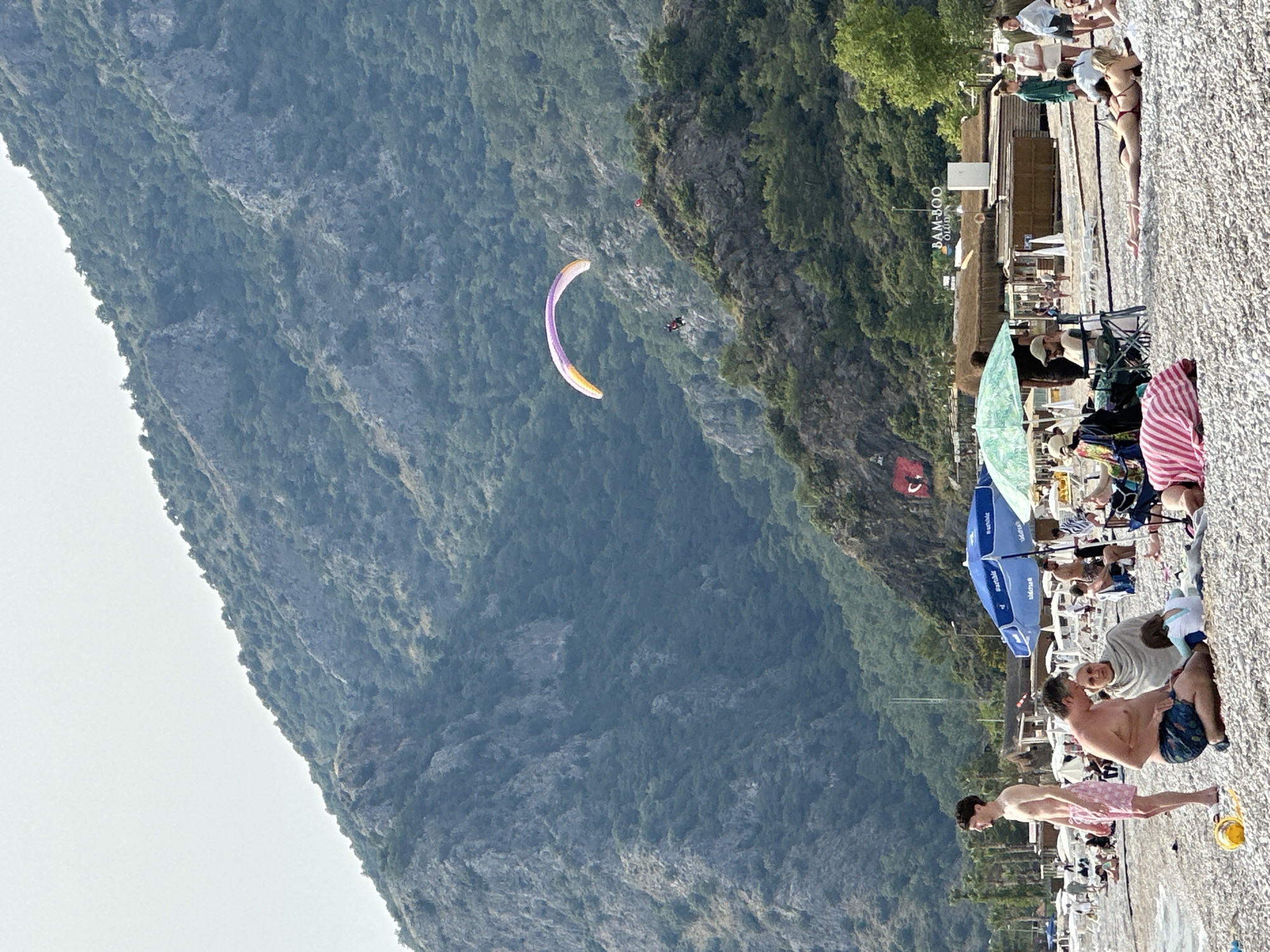 This photograph captures a scene at Ölüdeniz Beach in Fethiye, Turkey. The beach is populated with people sunbathing, swimming, and relaxing under umbrellas. Beachfront establishments, including the "Bamboo" bar, line the shore. In the sky, a paraglider soars against a backdrop of a towering mountain.