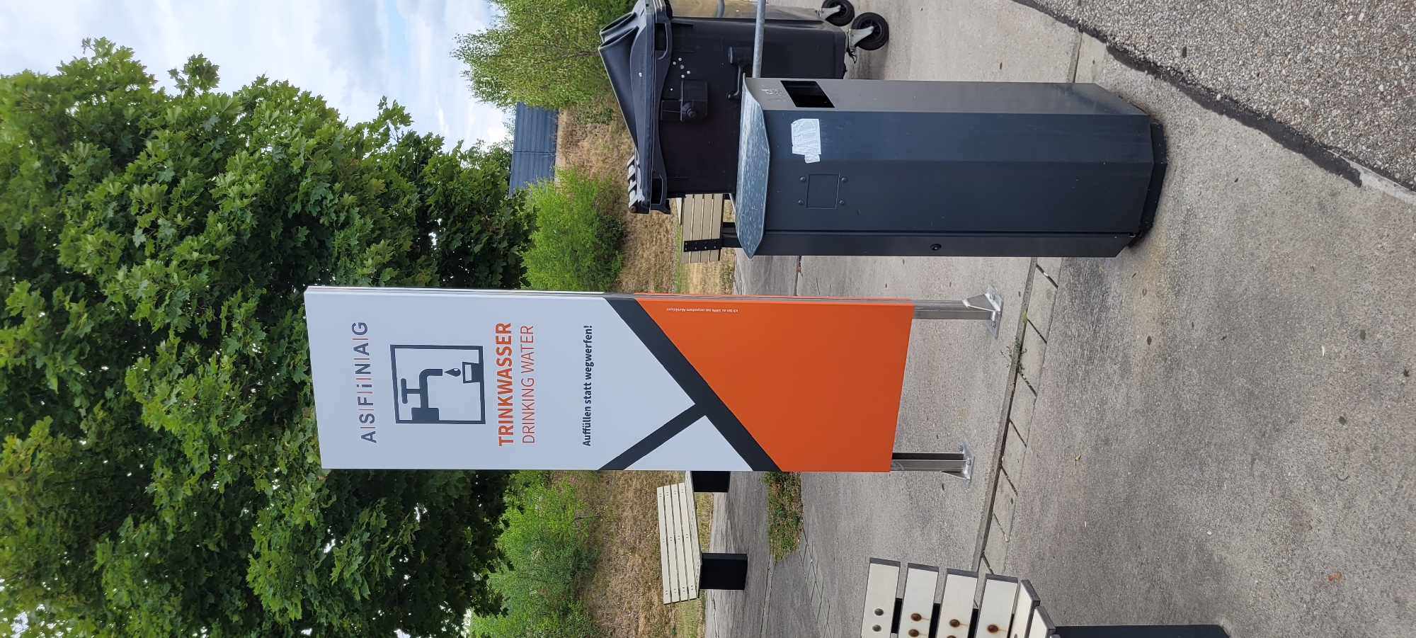 The image depicts an outdoor scene in Pöchlarn, Austria, near the Danube River. In the foreground, a dark gray, hexagonal trash receptacle stands on a concrete surface. To the left, a light-colored bench with a slatted seat and black legs is visible.
Dominating the center of the image is a tall, white sign with an orange and black triangular graphic at the bottom. The top of the sign features text and a symbol: "A|S|F|i|i|N|A|G" is written across the top, followed by a black outline of a faucet with a droplet of water falling into a cup. Below this, the text "TRINKWASSER" (German for drinking water) is in orange, and "DRINKING WATER" is in smaller black text. The phrase "Auffüllen statt wegwerfen!" (Refill instead of throw away!) is also present. A small inscription in the lower right corner of the orange section reads "Ich bin zu 100% aus recyceltem Aluminium!" (I am 100% recycled aluminum!).
In the background, another bench is partially visible, and a large, black wheeled trash bin stands to the right of the sign. Trees with lush green foliage form a backdrop, and a gray fence or barrier can be seen in the distance. The ground is paved with concrete slabs and gravel. The sky is overcast with scattered clouds.