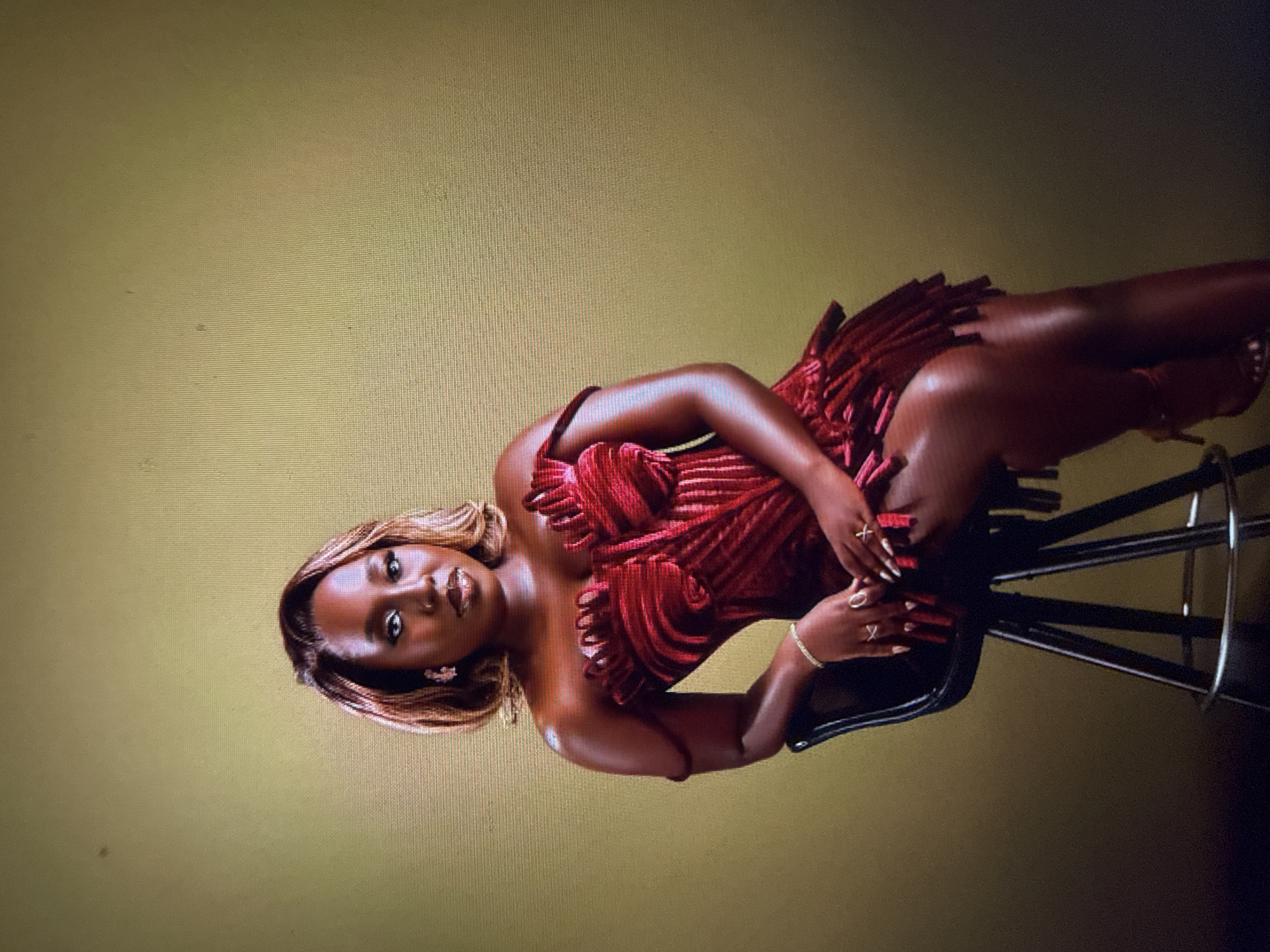 A woman with brown hair and tan skin sits on a black stool against a muted gold background. She is wearing a red strapless dress with a textured design. Her left arm is resting on the back of the stool, and her right hand is in her lap. She is wearing rings on her fingers and a bracelet on her left wrist. Her legs are crossed, and she is wearing high-heeled shoes. The scene is lit with soft, directional lighting, creating shadows on her skin and dress. There is no indication of the city of Abuja, Nigeria, or any other specific location in the image.