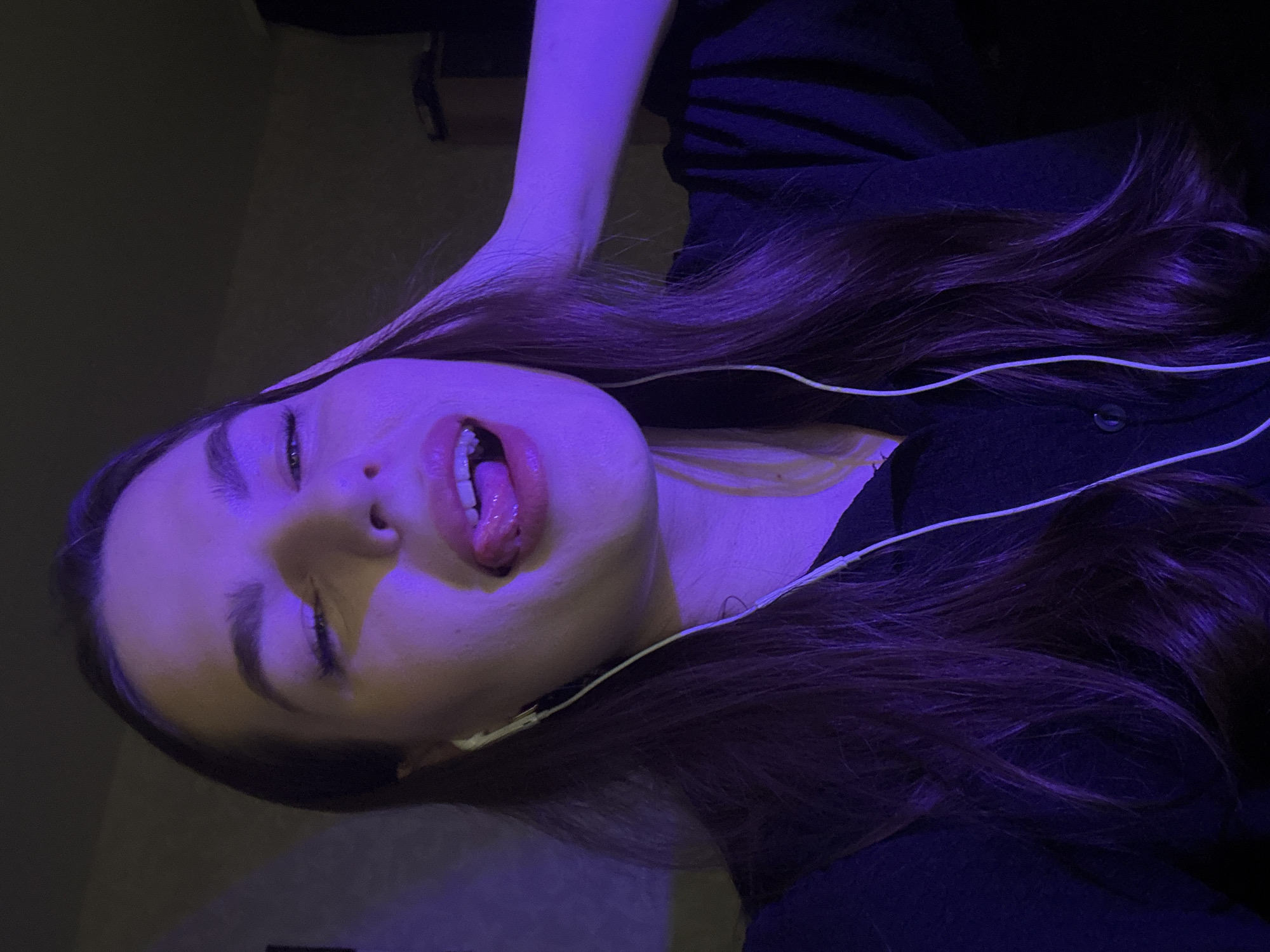 A young woman is lying on her back with her eyes partially closed and her tongue sticking out. She has long, dark hair and is wearing earphones. The lighting in the room is predominantly blue. The location is unknown, but the prompt suggests a connection to the city of Chaikovsky, Russia, which is unconfirmed by the image itself.