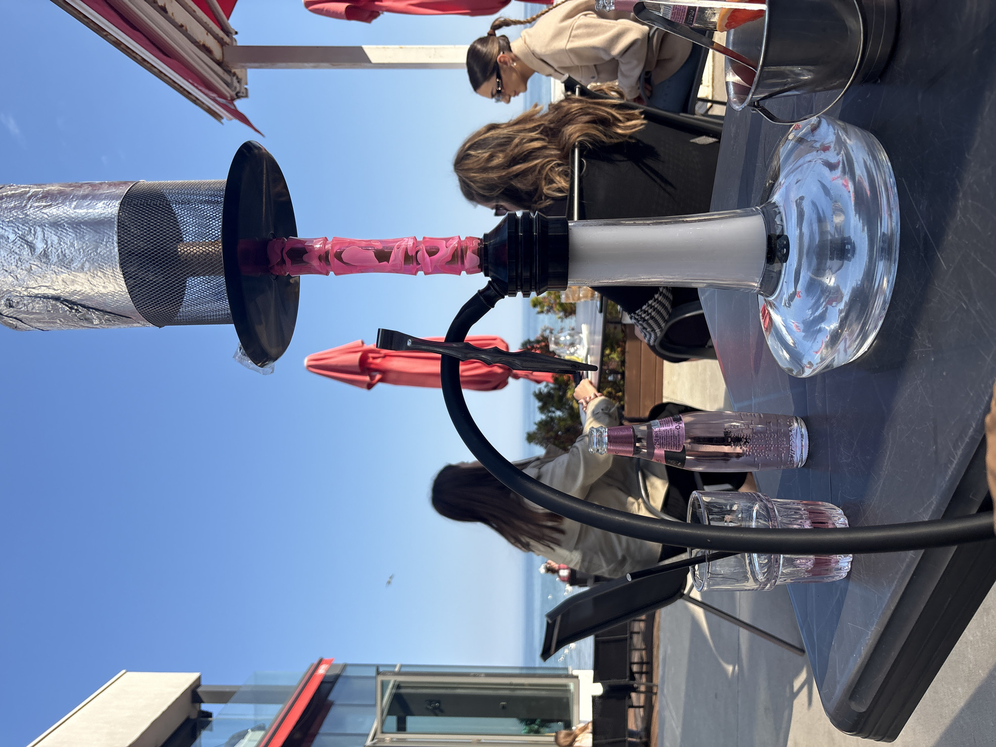 The image shows a hookah on a table outdoors. There are three people seated behind the hookah. In the background is a building with a red trim and glass windows. In the distance, the blue of the Black Sea contrasts with the clear sky. The image appears to have been taken in Varna, Bulgaria, based on the coastal location and architecture. There is also a bottle of pink liquid and a glass on the table.