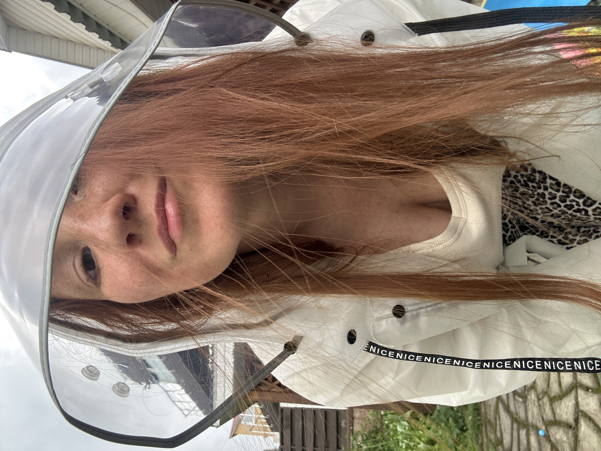A woman with red hair wearing a clear plastic visor and a white jacket is facing the camera. The jacket has the word "NICE" repeated along the band. The background includes a house, green foliage, and a brick-paved area. The location is not identifiable as Nizhny Novgorod, Russia from the image alone.