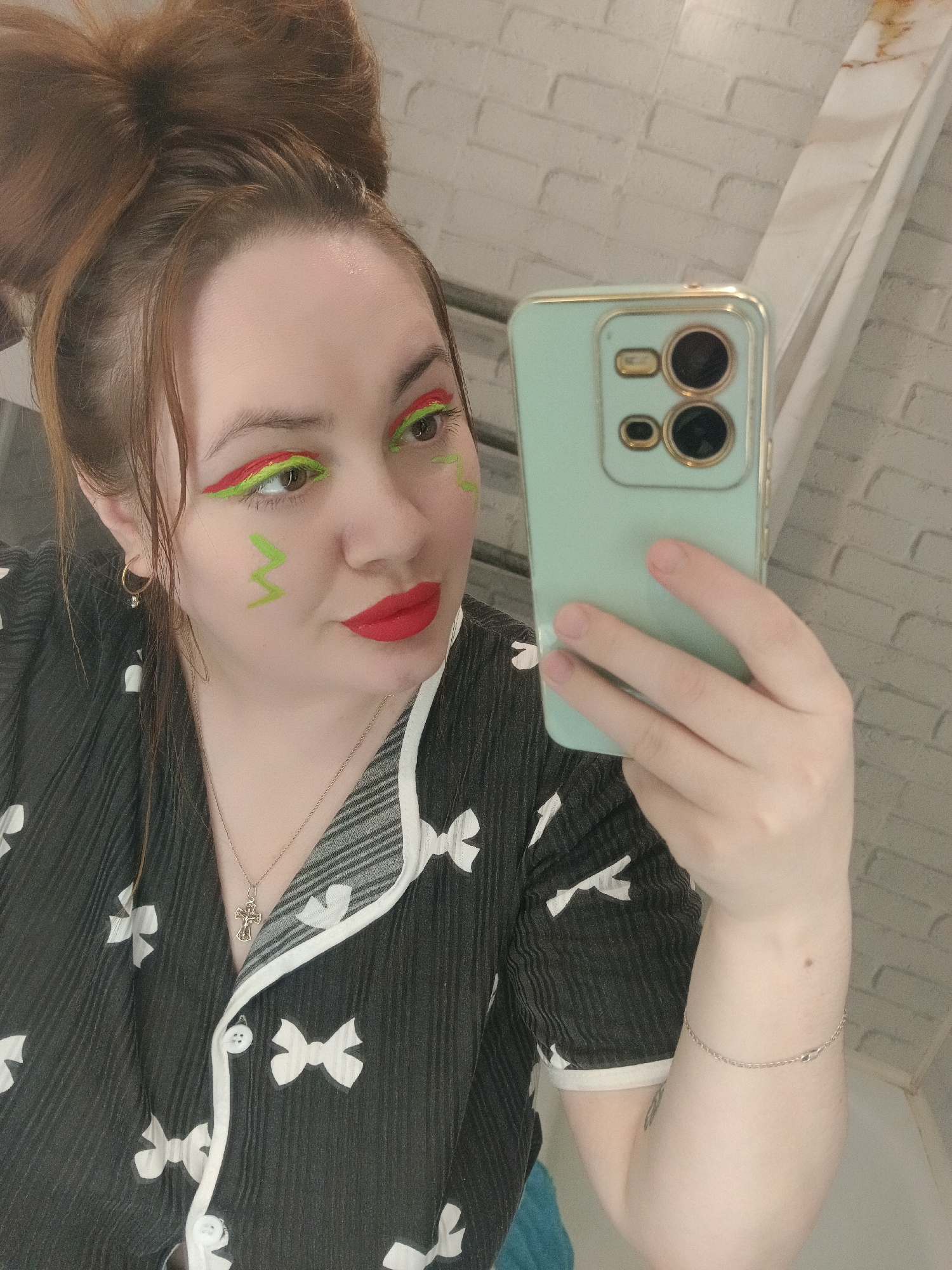 A medium close-up selfie features a woman with light skin and reddish-brown hair tied in a high bun. She is holding a light green smartphone with a clear case and gold-colored camera rings in her right hand, taking the picture. Her makeup is a notable feature, with bold red eyeliner above bright neon green eyeliner on both eyes, and bright red lipstick. Green zig-zag lines are painted on her left cheek.
She is wearing a black top or pajama shirt with a textured fabric and a pattern of white bows, featuring white trim along the collar and buttons. A gold-colored chain with a cross pendant is visible around her neck, and a small gold hoop earring with a charm hangs from her left ear. A thin silver chain bracelet adorns her right wrist.
Her expression is neutral, with her gaze directed slightly away from the phone's lens. The background consists of white, possibly brick-patterned, tiled walls and a white vertical fixture with a light-colored marble-like top, suggesting an indoor setting like a bathroom or dressing room. A glimpse of blue fabric is visible in the lower left of the frame. This scene takes place in Chaikovsky, Russia.
