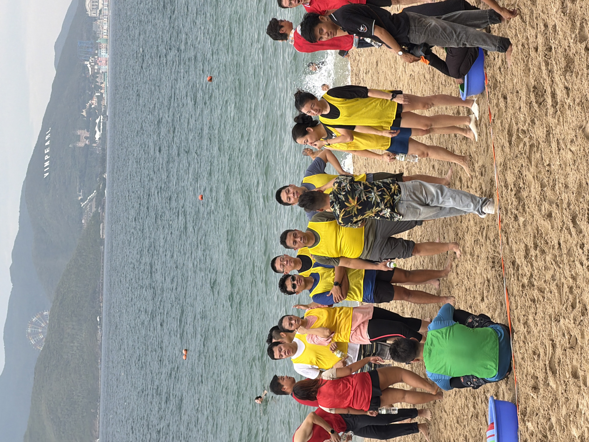 In Nha Trang, Vietnam, a group of approximately 15-20 people are gathered on a sandy beach with the ocean in the background. The water is choppy, and several orange buoys are visible. In the distance, a hillside is covered in green foliage, with the white letters "VINPEARL" prominently displayed near the summit, along with buildings and a Ferris wheel.
The individuals are diverse in age and gender. Many are wearing brightly colored athletic vests in yellow and red, suggesting a team activity or event. Some are holding plastic water bottles. One person is seated on the sand facing away from the camera, wearing a green vest. Another person is partially submerged in a blue plastic tub, with their head and upper body inside. The mood appears casual and possibly related to a beach activity or competition. The overall environment is a coastal setting with a clear view of the ocean and the mountainous landscape beyond the city.
