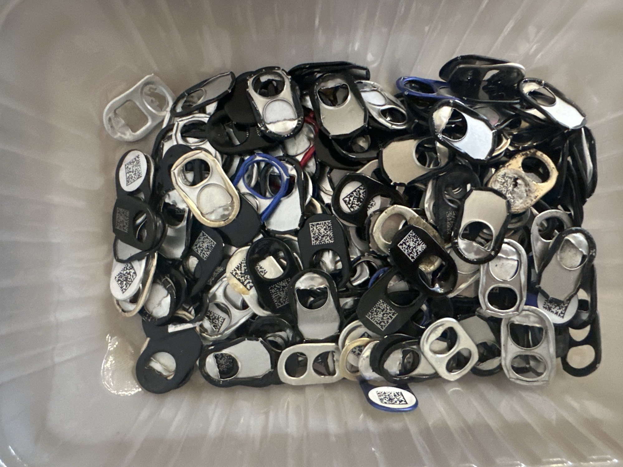 The image shows a pile of can tabs, many of which have QR codes printed on them. They are contained within a translucent plastic bag or bin, which appears to be in a light-colored environment, possibly a room or storage area. The can tabs are varied in color, with many being silver and black, and some appear to have blue or red elements, possibly from rubber bands or other materials. Some tabs are layered on top of each other, creating a dense pile. There is a small amount of a translucent, viscous substance near the top left of the image.
There is no information in the image to suggest a location in Nevinnomyssk, Russia. The scene is devoid of people or any identifiable human activity. The focus is entirely on the collection of can tabs.
