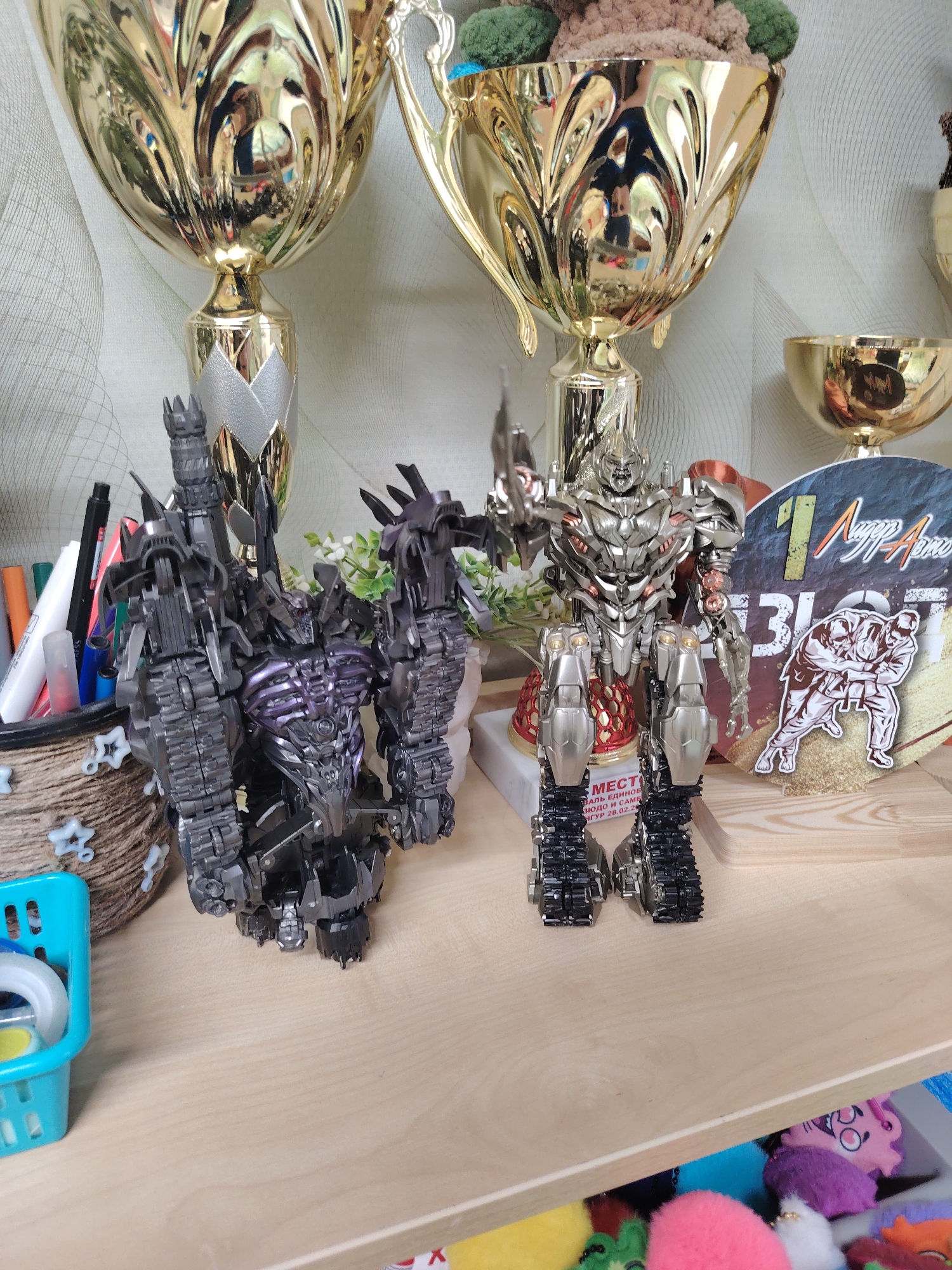 The image depicts a shelf in what appears to be a private residence. Located on the shelf are two Transformers toys: one is predominantly black and purple with a tower-like structure on its left arm and a spiky appendage on its right, the other is silver with a blade in its right hand. Behind them are several gold trophies. A pen holder filled with pens and markers sits to the left of the black transformer. There's also a blue plastic basket with tape and other small items. To the right is a wooden cutout displaying two figures wrestling and text written in Russian that includes the number '1' and the word "Blot". The wrestling cutout also includes the text "MECTO. АЛЬ ЕДИНОБ..." and the text "...ЮДО И САМБ... ГУР 28.02.28...". The background is a light wallpaper with a subtle pattern. Beneath the shelf, a drawer is partially open, revealing colorful plush toys. Based on the text written on the wrestling cutout, it is likely that the image was taken in Chaikovsky, Russia.