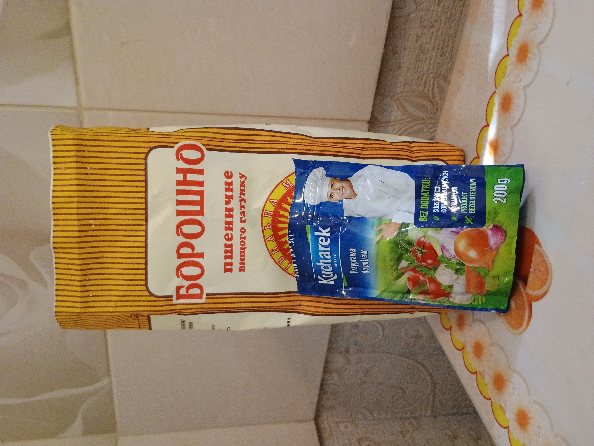 This is a close-up, top-down shot of a bag of flour and a small packet of seasoning, possibly on a kitchen counter. The bag of flour, labeled in Cyrillic script, appears to be "Борошно" (flour) and specifies "пшеничне вищого гатунку" (wheat flour of the highest grade). The seasoning packet, labeled "Kucharek Przyprawa do potraw" (Kucharek seasoning for dishes) in Polish, advertises "BEZ DODATKU: SUBSTANCJI KONSERWUJĄCYCH, AROMATÓW" (without added preservatives or flavorings) and "PRODUKT BEZGLUTENOWY" (gluten-free product). The packet also shows a net weight of 200g and features an image of a chef. The overall color palette is warm, with yellow and brown tones from the flour bag and the vibrant colors of the seasoning packet. The background suggests a home kitchen setting, with tiled walls and a patterned tablecloth. The lighting is natural, indicating it was taken during the daytime. The image is static and does not depict any specific event or emotion.