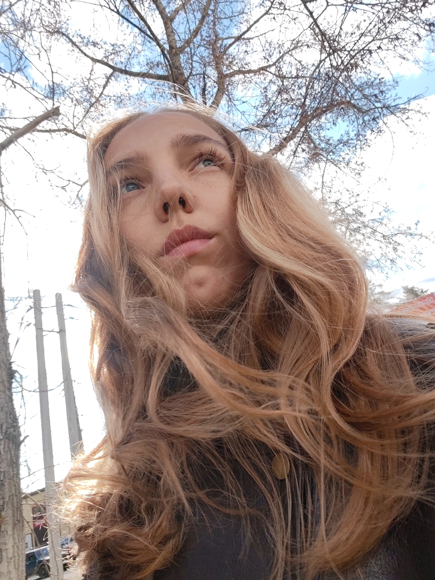 A low-angle, eye-level, medium close-up shot depicts a young woman with long, light brown wavy hair. The subject’s face is slightly tilted upwards and to the left, and her gaze is directed toward the upper left of the frame. She has fair skin, light eyes, and a nose ring. She wears a dark-colored jacket with a visible button near the lower right edge of the frame.
The background includes a tree with bare branches against a partially cloudy, bright sky. Several utility poles and the top of a tree are visible behind the subject. The lower left corner shows a tree trunk and a glimpse of a building and parked cars.
The location is recognizable as the city of Chaikovsky, Russia, because of the architectural style of the buildings visible in the background, and the types of trees present in the scene.