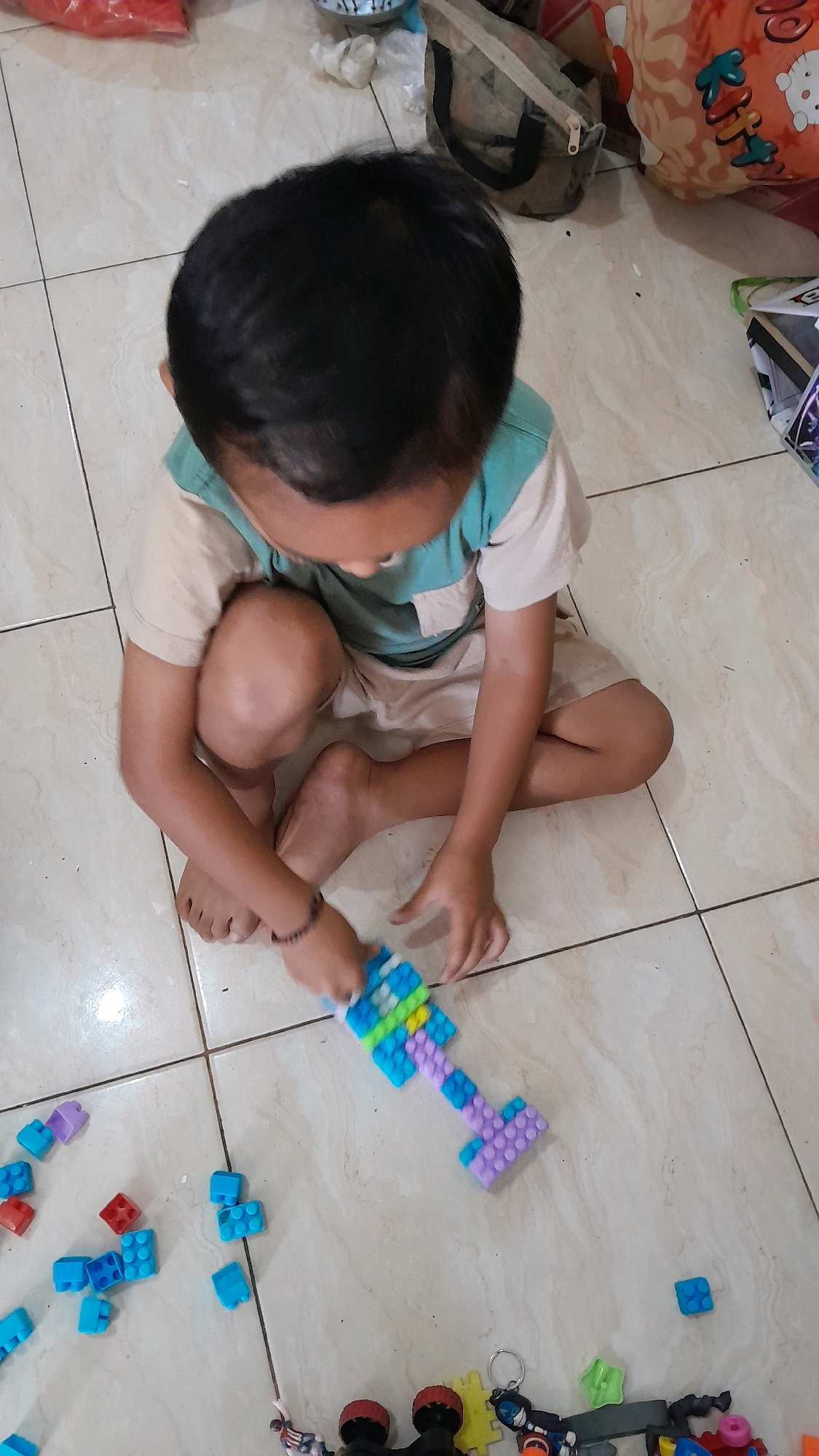 A young boy with black hair, wearing a tan and teal t-shirt and tan shorts, is sitting on a tiled floor, playing with colorful building blocks. The blocks are scattered around him and he is holding a completed structure resembling a key. The floor appears to be indoors, and the lighting suggests it is daytime. In the background, there are various bags and some colorful items on the floor.