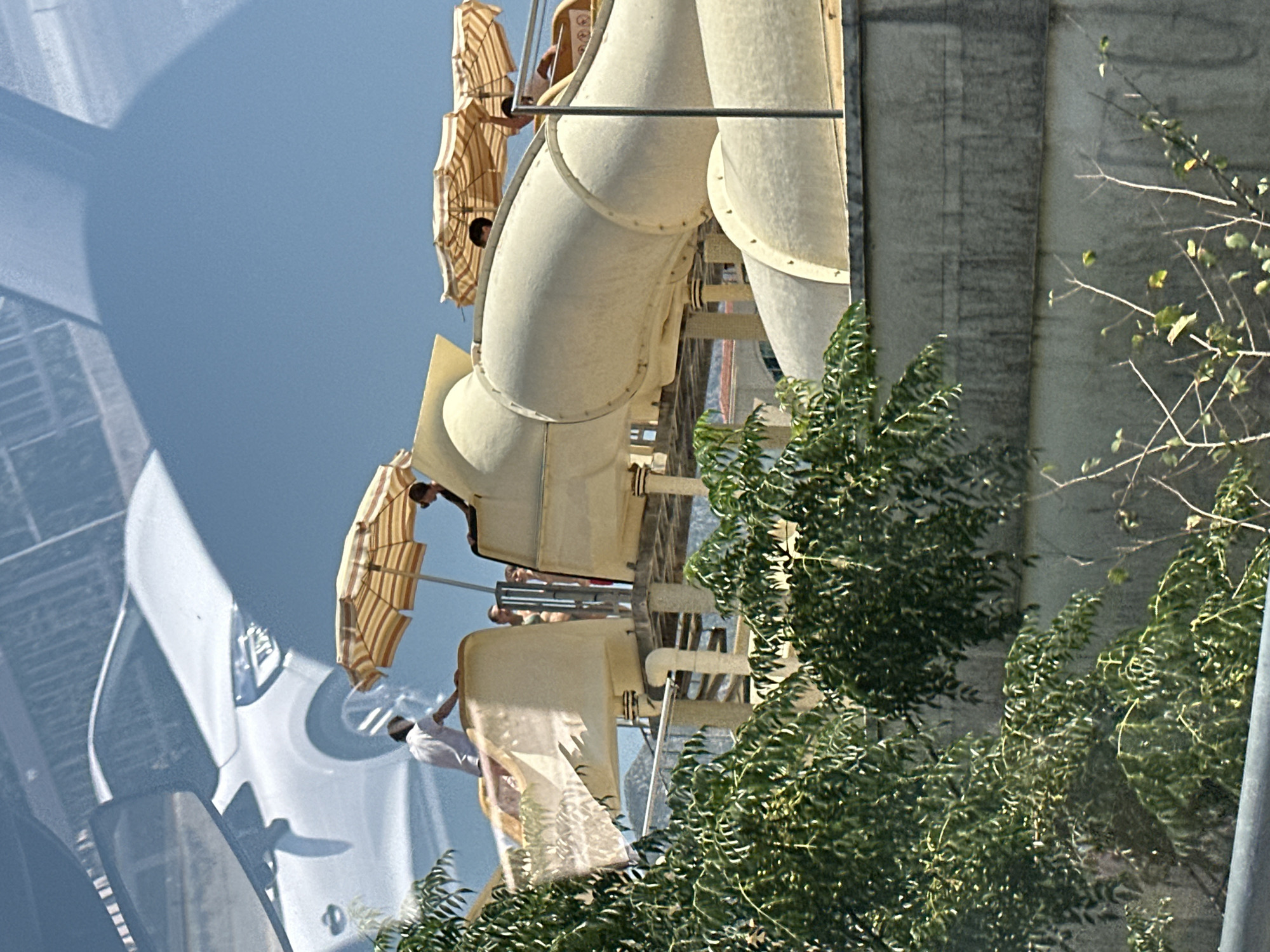 The image depicts a scene at a water park, likely in Boreti, Montenegro. In the foreground, lush green trees obscure a portion of the view. The main subject is a water slide structure, constructed of a light-colored, possibly fiberglass material. Several individuals are visible on and around the slide. A person in a white shirt stands at the top of a section of the slide, appearing to prepare for a descent. Two umbrellas with a striped pattern of yellow and white are positioned over parts of the slide area, providing shade. One person is seated near one of the umbrellas. In the background, a white car is visible, reflected on a smooth surface, possibly a window or a shiny building facade. The sky is a clear blue. The water slide appears to be elevated, with support structures visible underneath. A concrete wall runs along the lower part of the image, separating the viewer's vantage point from the water park.