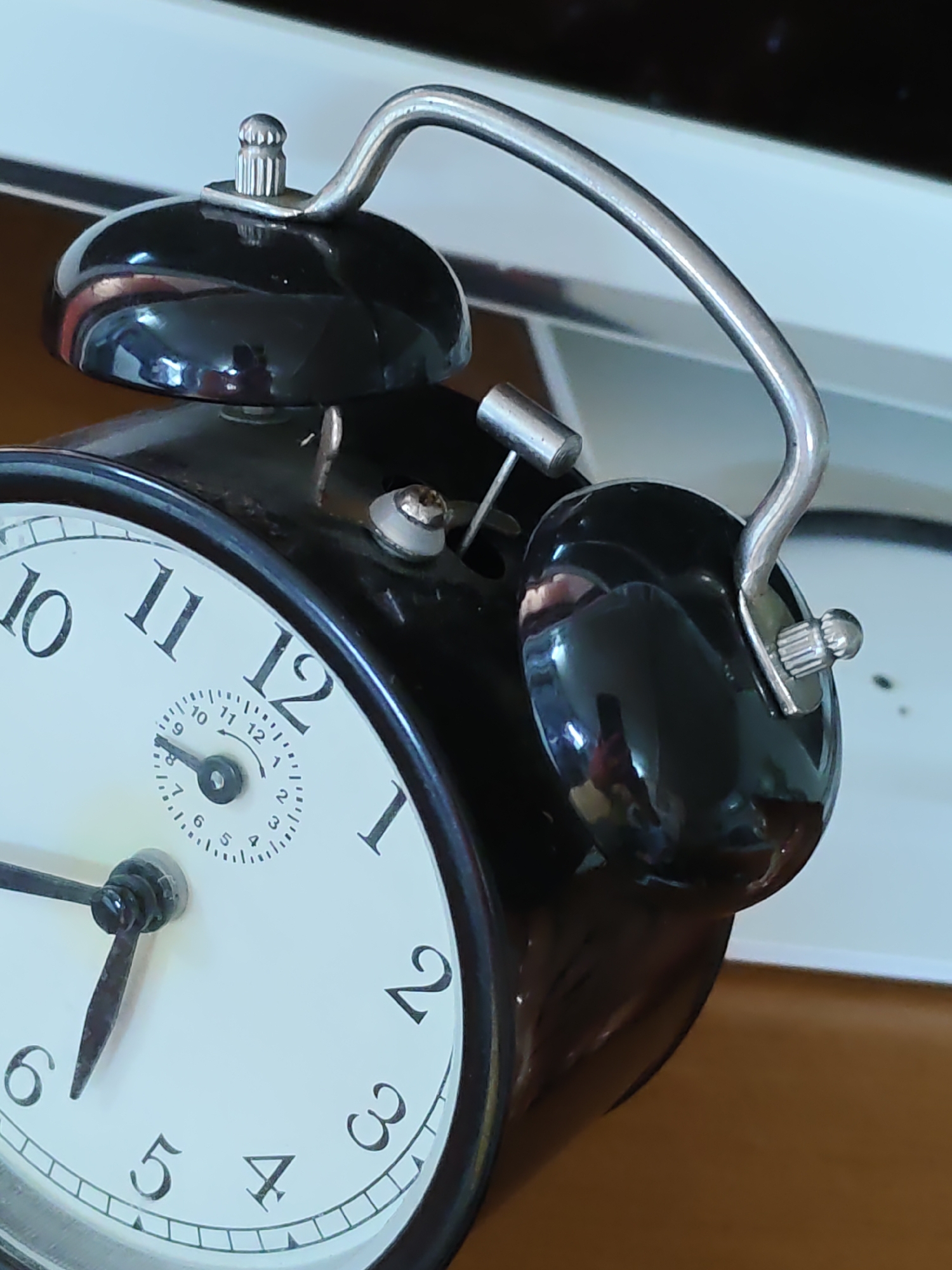 An extreme close-up shot of a black, vintage-style alarm clock is presented. The clock features a white face with black Arabic numerals and hands. The hour hand points just before the 6, and the minute hand points to the 4, indicating the time is approximately 5:20. There's a smaller dial within the main face, displaying numbers 1 through 12 for an unknown purpose. The clock has two bells on top, connected by a silver metal arch. The surface of the clock is highly reflective, showing distorted shapes and colors from its surroundings. The background is out of focus, appearing as a mix of white and muted colors, suggestive of an indoor environment. There is no indication of the city of Olginskaya, Russia, or any people or actions occurring.
