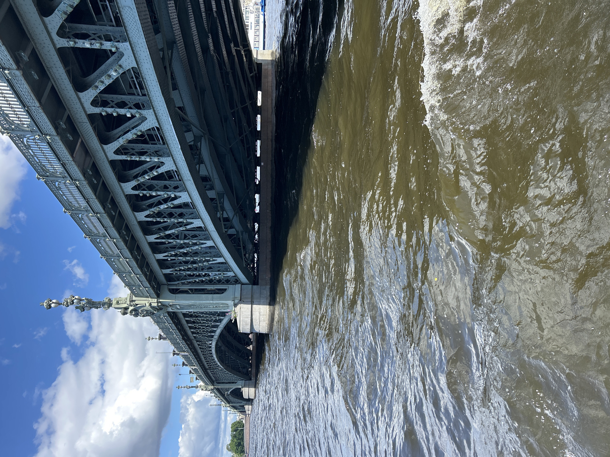 This is a low-angle, wide shot of a large, metallic bridge seen from below, with a body of water in the foreground. The bridge is intricate, with a complex network of girders and supports, and ornate lampposts attached to its side. The water is choppy and appears to be flowing, creating white foam on the right side of the frame, suggesting movement, perhaps from a boat passing by or the wake of a vessel. The sky above the bridge is blue with scattered white clouds. The overall scene is industrial and architectural, focusing on the scale and design of the bridge structure. There is no visible text in the image. The lighting suggests it is daytime, and the presence of clouds indicates mild weather. The angle of the shot and the focus on the underside of the bridge create a sense of looking up at a massive structure.