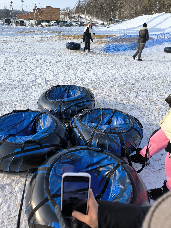 The image depicts a winter scene at a snow tubing facility in Prybuzke, Ukraine. In the immediate foreground, four black and blue snow tubes are visible, arranged on packed snow. A person's hands, gloved in black, hold a white smartphone positioned over one of the tubes, capturing a horizontal image of a snowy landscape. To the right of the foreground, the lower torso and legs of another person are visible, wearing bright pink snow pants and black boots, standing near the snow tubes.
In the mid-ground, a child in dark winter clothing pulls a single black and blue snow tube across the snow-covered ground. An adult in a white jacket stands near the child. Further to the right, another adult, dressed in a dark jacket and black hat, walks across the snowy terrain, moving past a section of blue safety netting or fencing. Another snow tube is visible near this walking adult.
The background features a snow-covered hill or slope on the right. To the left, several multi-story brick-colored buildings are visible, beyond which a parking area contains numerous parked cars and SUVs. Utility poles with overhead lines are present. The overall environment is bright, suggesting daytime conditions with a clear sky, and the ground is consistently covered in snow.