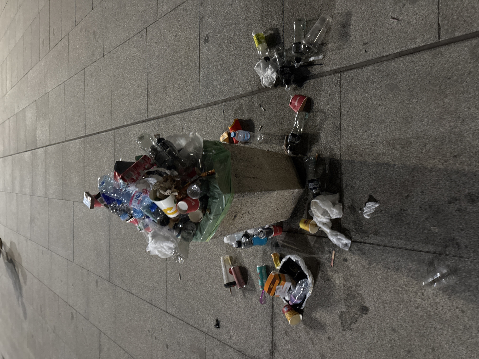 The image depicts an outdoor urban scene, likely a public pedestrian area or sidewalk, characterized by an overflowing waste bin and substantial litter scattered across the paved ground. The specific location within Sofia, Bulgaria, cannot be definitively identified from the visual information provided.
At the center, a light-colored, possibly concrete, public waste bin is visible, its top portion revealing a green plastic liner. The bin is severely overfilled, with a large mound of refuse extending significantly above its rim. This refuse consists of various items, including numerous plastic bottles (clear and colored, such as blue), several aluminum beverage cans (some red, possibly Coca-Cola branded), white and yellow paper cups (one with a partial McDonald's logo), and crumpled plastic bags and wrappers.
The ground surrounding the bin is paved with rectangular grey concrete or stone slabs. This pavement is extensively littered. To the immediate left and front of the bin, items include a clear plastic bottle, a small white plastic bag containing an orange cardboard box (resembling a burger container) and other debris, two small colored metal cans (one pinkish-red, one teal-blue), a large transparent tube-like object (possibly a disposable vape or syringe without a needle), and several cigarette butts.
To the right and front of the bin, more litter is present. This includes multiple clear glass or plastic bottles (some appearing broken or empty), some with red caps or labels, a yellow plastic bottle, empty yellow and red paper cups, and various plastic wrappers and small dark objects, possibly electronic waste or parts. Several small, reflective wet patches are discernible on the pavement, particularly towards the bottom of the image, possibly indicating spilled liquids or moisture.
The scene is illuminated, suggesting either artificial lighting at night or dusk, or an overcast day. No identifiable people are visible, though a blurred shadow in the upper left corner might suggest a passerby. The overall impression is one of significant urban waste accumulation in a public space.