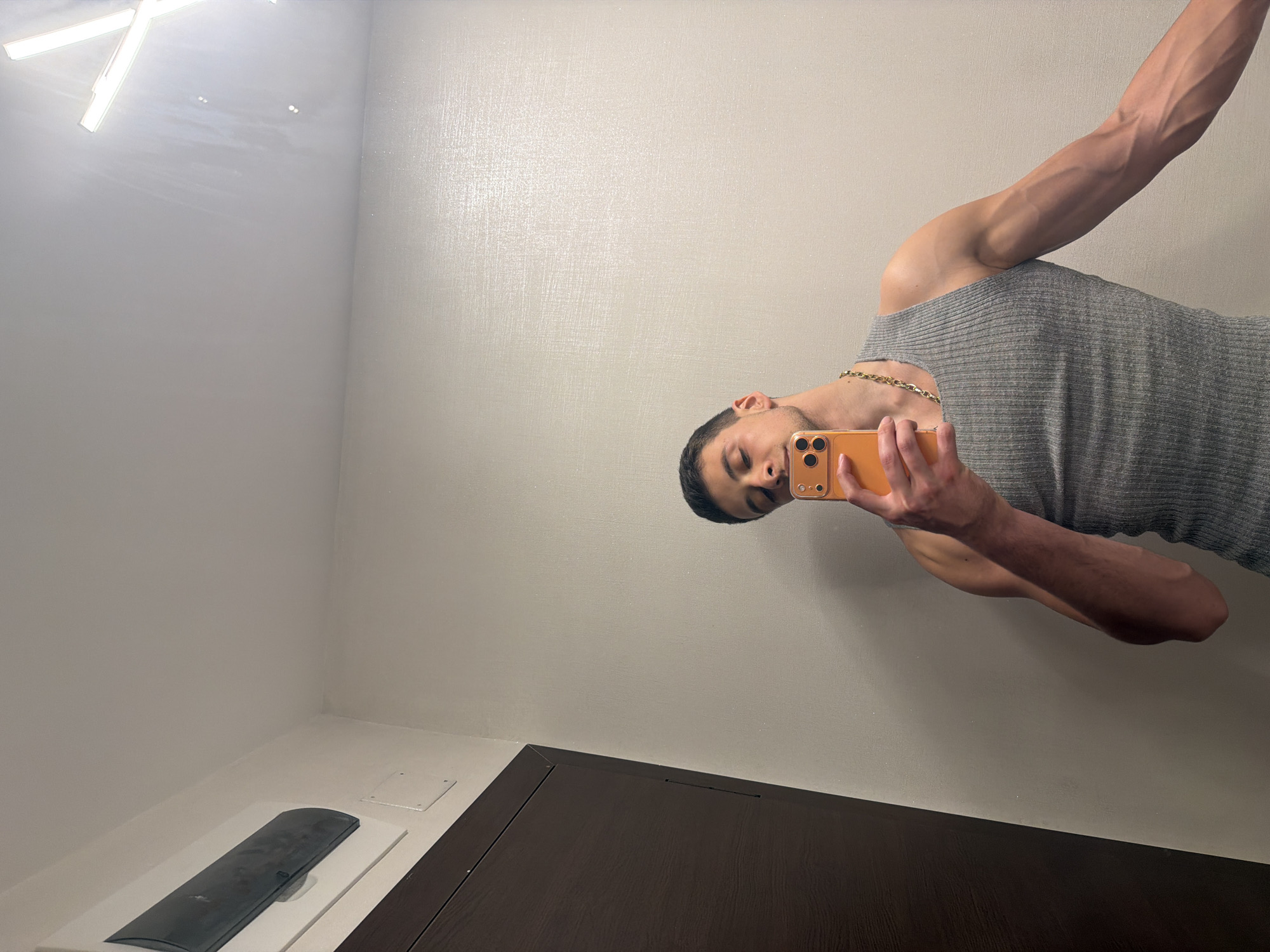 The image depicts a mirror selfie taken by a person in an indoor setting. The photo is rotated 90 degrees counter-clockwise.
**Subjects:** The primary subject is a person, likely male, with short dark hair, seen from the chest up. He is wearing a grey ribbed tank top and a gold-colored, possibly Cuban link style, chain around his neck. His right arm is prominently featured, showing muscular definition and visible veins. His face is turned slightly downward, eyes seemingly focused on the phone he is holding.
**Activity or event taking place:** The person is taking a mirror selfie using an orange-colored smartphone, which he holds in his right hand.
**Objects:** Apart from the person and his attire, a bright, rectangular light fixture is visible on the ceiling in the upper left. In the bottom left corner, there is a white counter or shelf with a black, elongated object (possibly a remote control or an electronic device) resting on it, next to a white square cover. A dark brown wooden surface, possibly a cabinet or vanity, extends across the bottom portion of the image.
**Scene, setting, and notable details:** The setting appears to be an interior space, likely a bathroom or dressing area, given the presence of a mirror, counter, and overhead lighting. The walls are a light, textured off-white or beige color. The lighting is bright and artificial, casting reflections on the mirror. No specific time of day or weather can be determined. The person's expression is neutral, concentrating on taking the picture. No visible text is present in the image.