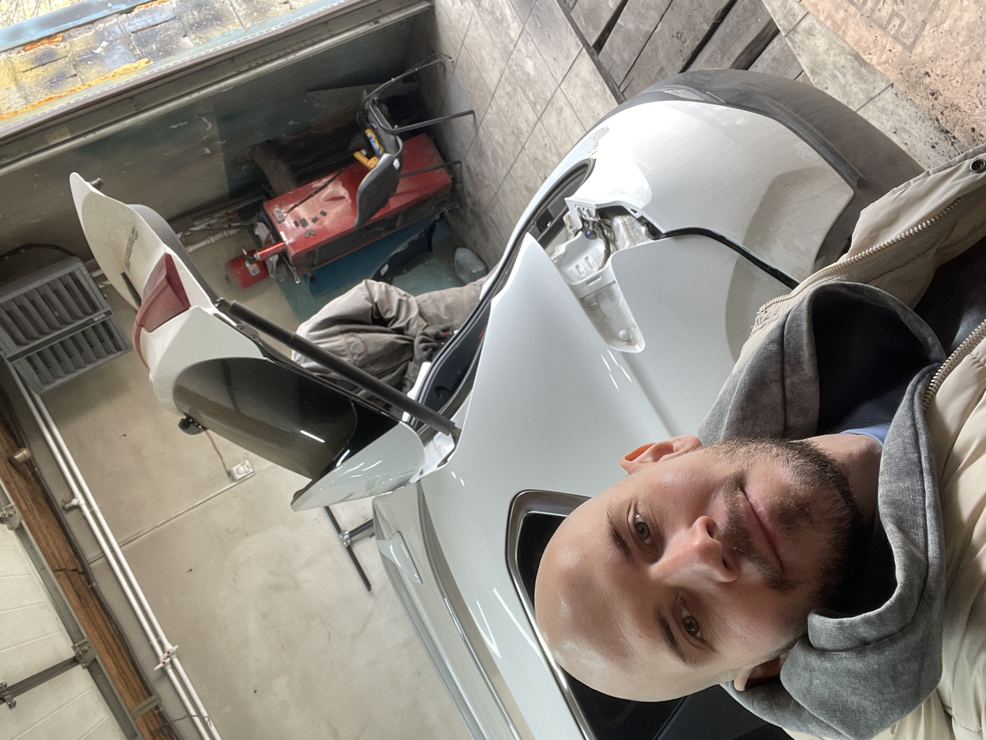 A man with a shaved head and a short beard, wearing a grey hoodie and a beige jacket, is taking a selfie in a garage or workshop. The subject is positioned near the front of a white car that appears to be undergoing repairs, with parts of the car disassembled, particularly around the front bumper and hood area. In the background, there is a red tool cart and assorted garage equipment. The location is Минино, Russia.