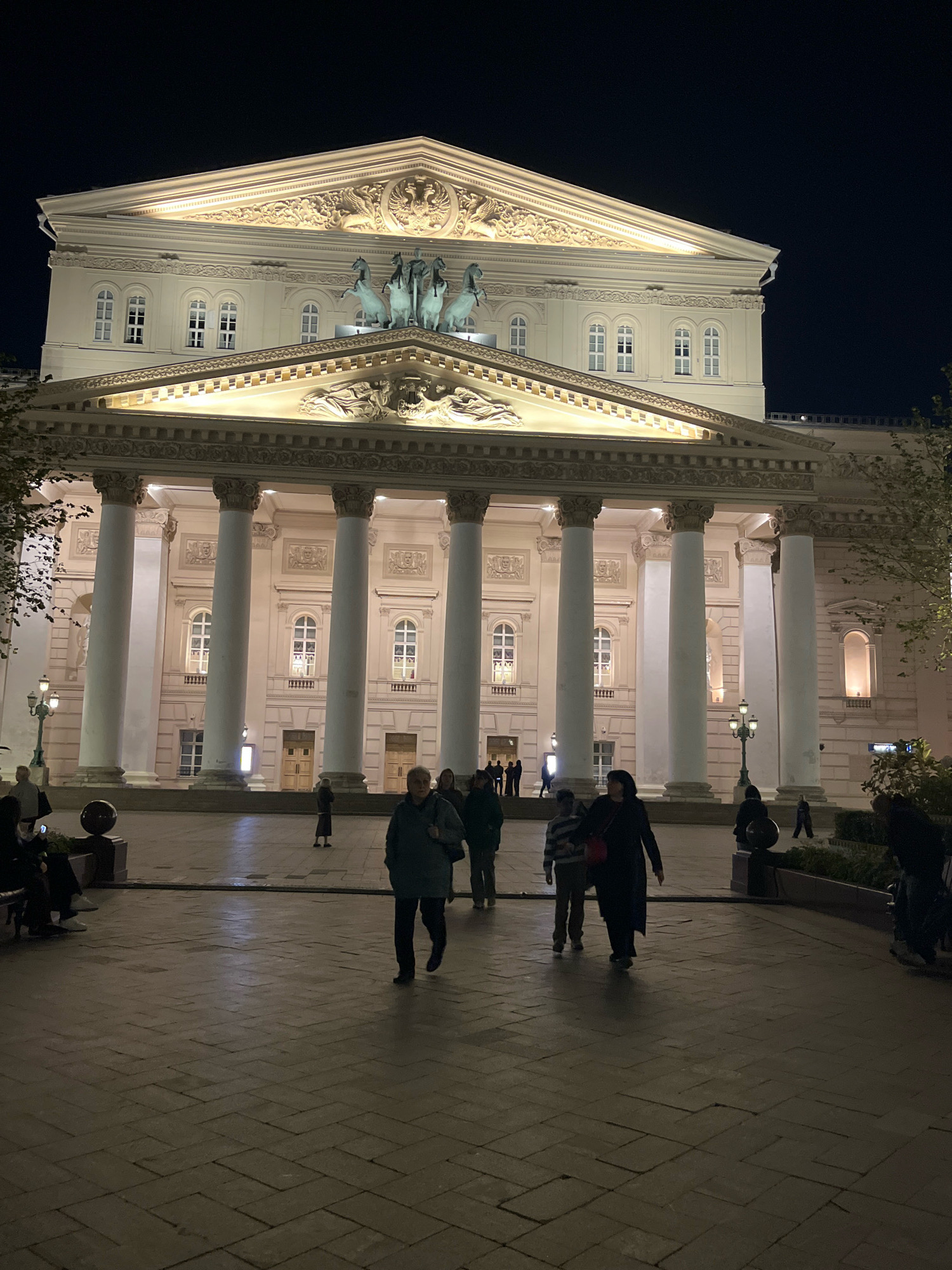 The image shows the Bolshoi Theatre in Moscow, Russia, at night. The grand neoclassical facade of the theatre is illuminated, highlighting its columns, ornate pediment, and sculptures. Several people are walking on the paved plaza in front of the theatre. The plaza is lit by lampposts and the theatre's exterior lighting, casting long shadows on the ground. Trees with sparse foliage are visible on the sides of the building. The sky is dark, indicating it is nighttime.