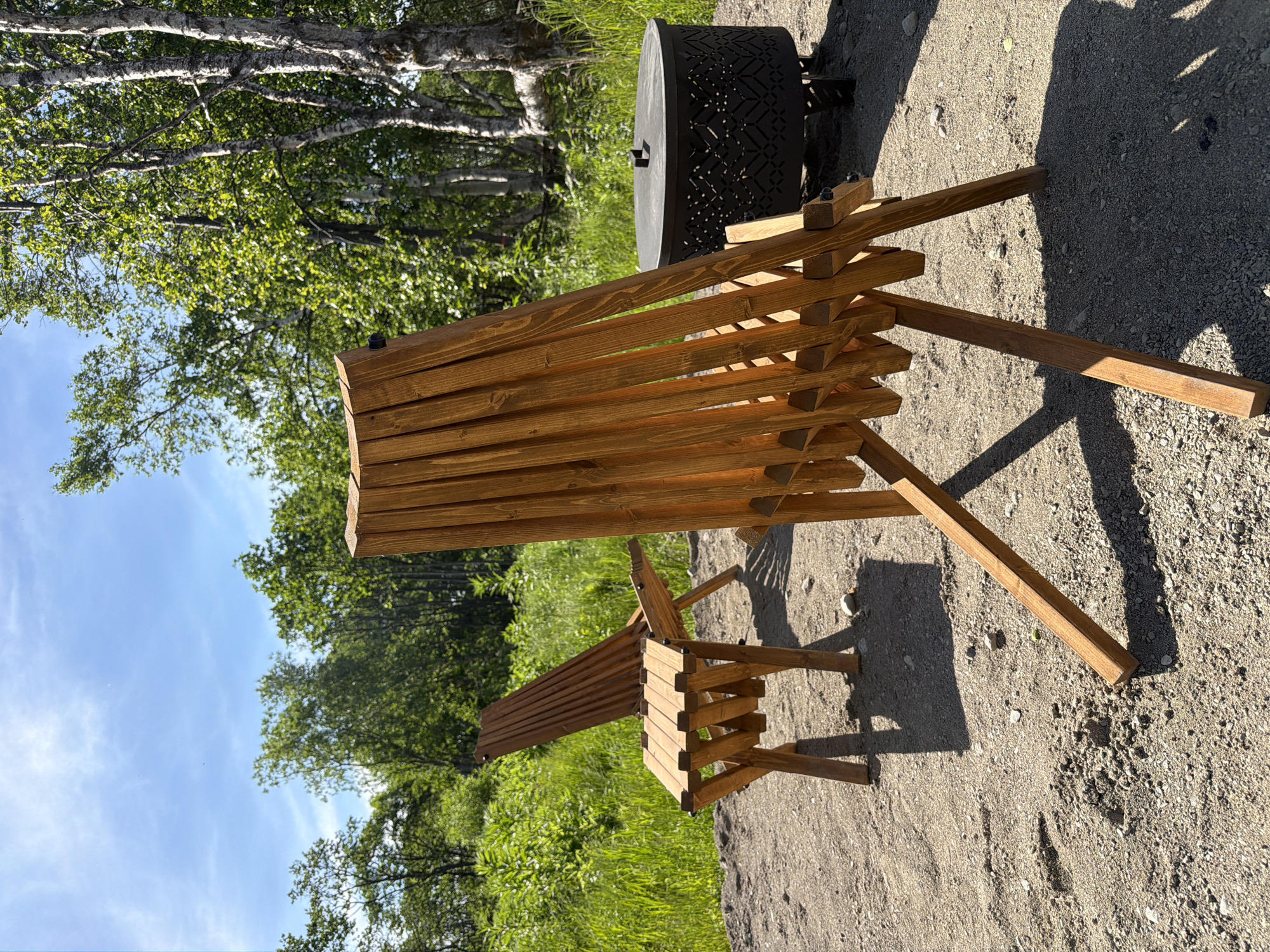 The image depicts an outdoor scene with two wooden chairs and a fire pit. One chair is in the foreground, facing away from the viewer, with its back to the camera. The other chair is positioned behind and to the left of the first chair, also facing away. Both chairs appear to be made of slatted wood with a natural finish. To the right of the chairs and slightly behind them is a dark, round fire pit with a decorative patterned exterior and a lid. The ground is covered in gravel and sand. In the background, lush green foliage and trees are visible, with a bright blue sky overhead. The lighting suggests it is daytime. There are no people present in the image. The scene appears to be a recreational area, possibly a backyard or a campsite, in a natural setting. The location "Новый, Russia" is not visually represented or implied by the image's content.