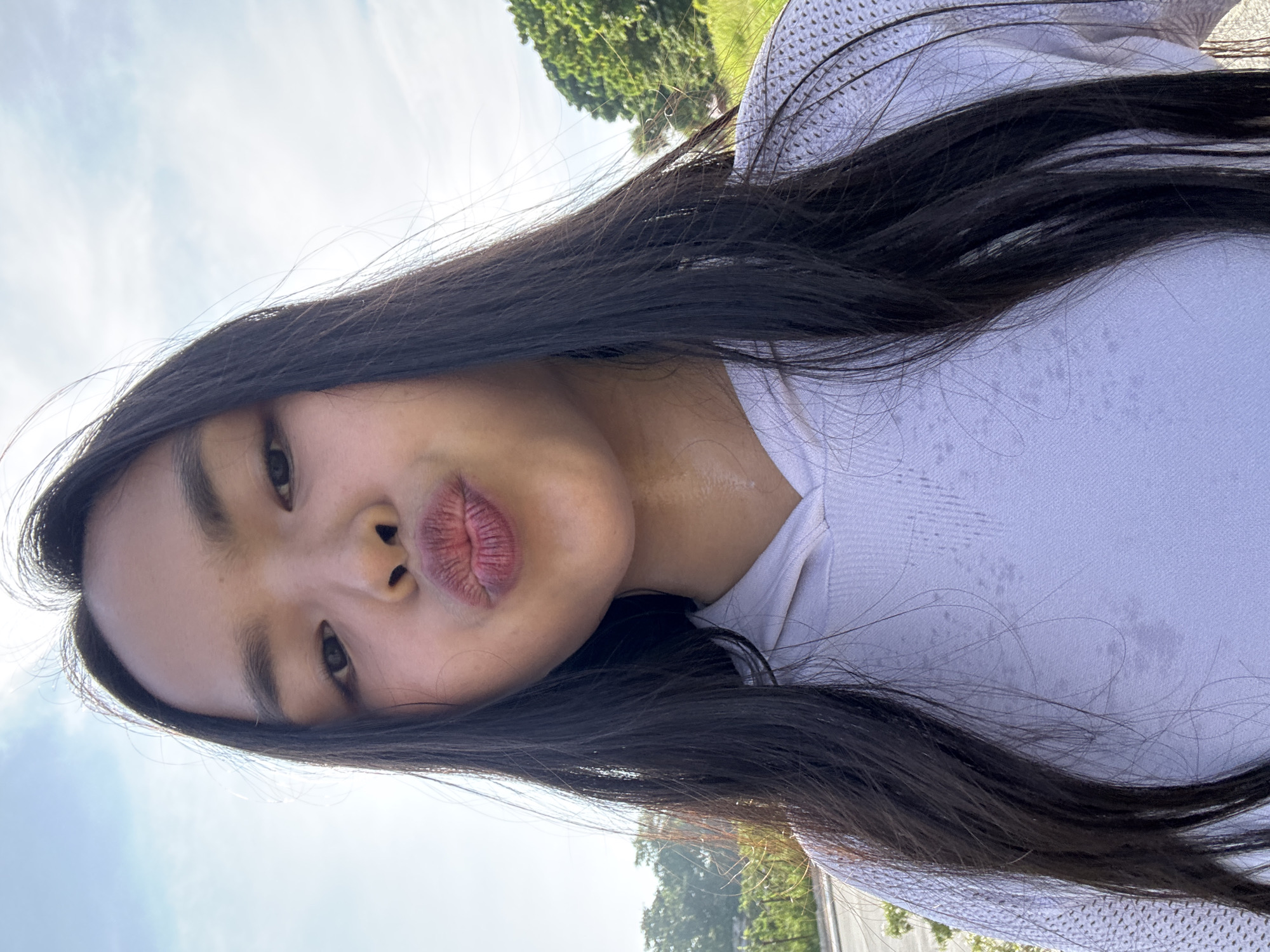 A young individual with long, dark hair is captured in a selfie outdoors, puckering their lips towards the camera. They are wearing a light-colored hooded sweatshirt. The background suggests a location in Davao City, Philippines, with hints of greenery, trees, and a partially visible road or pathway, possibly indicating a park or roadside setting. The sky above is bright with some clouds. The individual appears to be in good health.