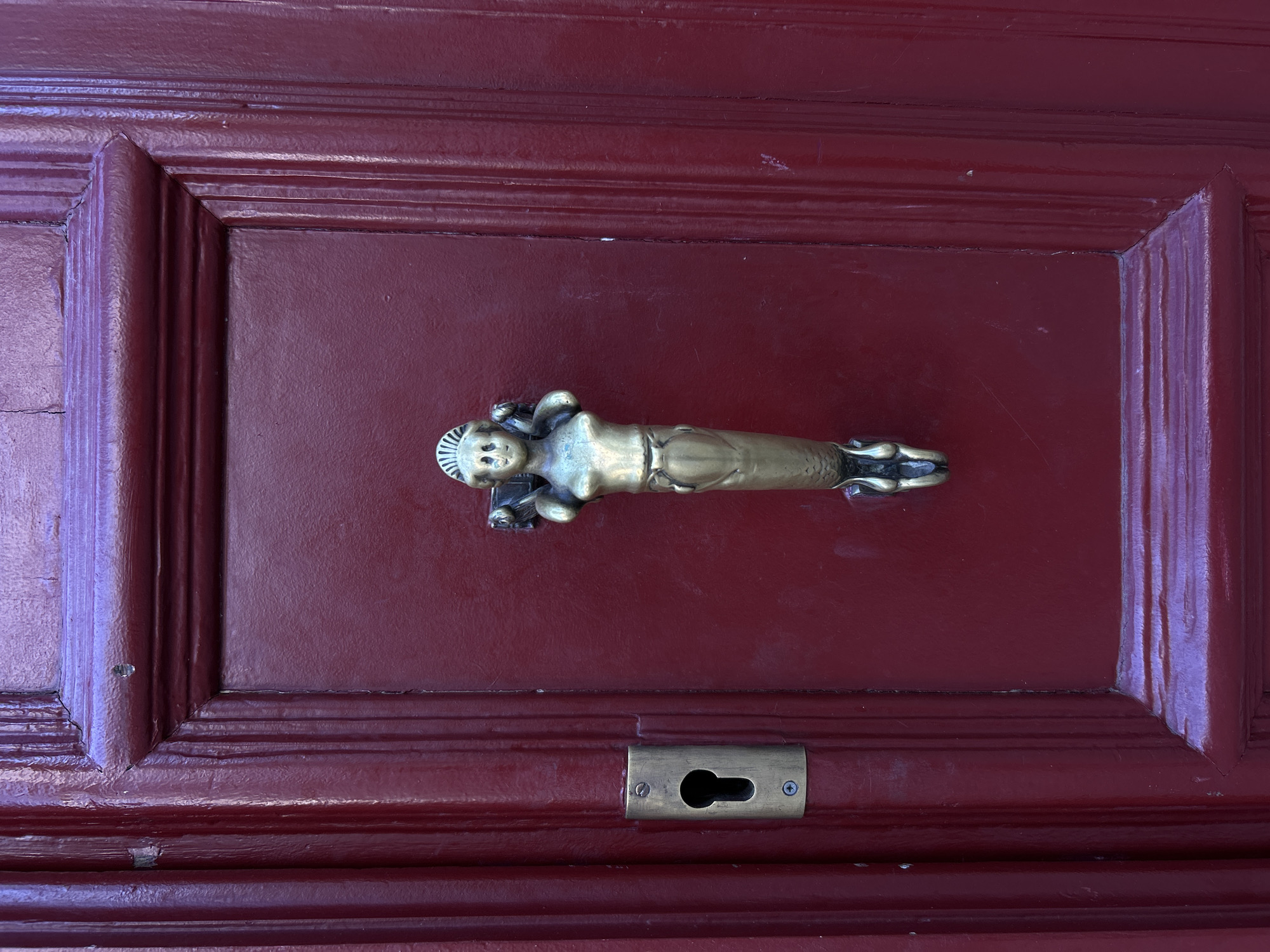 This image shows a close-up of a rich, dark red wooden door with ornate detailing. A brass door knocker, shaped like a mermaid with a human torso and a fish tail, is prominently featured in the center of the door. To the left of the knocker, a brass keyhole plate is visible. The wood of the door exhibits a high-gloss finish and features raised paneling. The overall impression is one of classic, somewhat antique, European architecture. The image was taken in Paris, France.