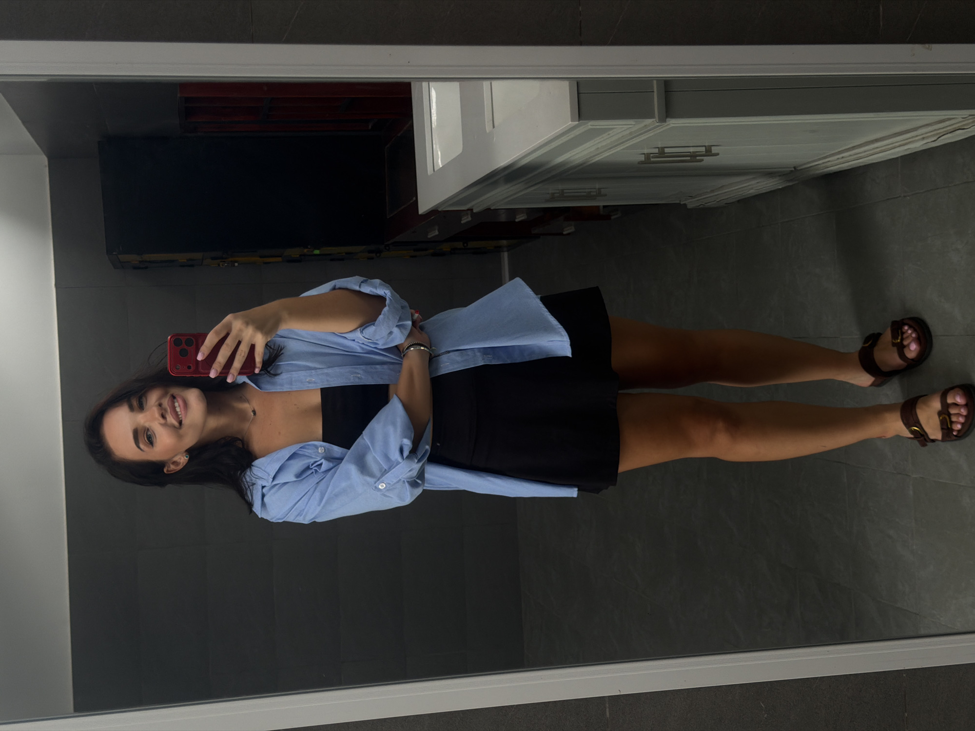 A young woman is taking a selfie in a bathroom. She is wearing a black top, a black skirt, and a light blue button-down shirt. She is holding a red phone in her right hand. The bathroom has gray tile flooring and a white vanity. The location is Bac Nha Trang Ward, Vietnam.