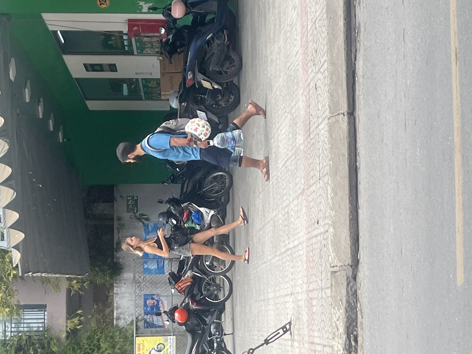 The image depicts a street scene in Đà Nẵng, Vietnam. On the right, a man with dark hair and a beard, wearing a blue t-shirt and dark shorts, walks on the sidewalk. He carries a backpack and holds a large water bottle and a decorative object in his hand. To his left, a woman with blonde hair, wearing a black tank top and denim shorts, walks in the same direction on the sidewalk. Behind them, several parked motorcycles are visible, lining the street. The environment includes a building with a green facade and large windows. The sidewalk is paved with patterned tiles and has a concrete curb. The street in the foreground is asphalt with a yellow line marking.