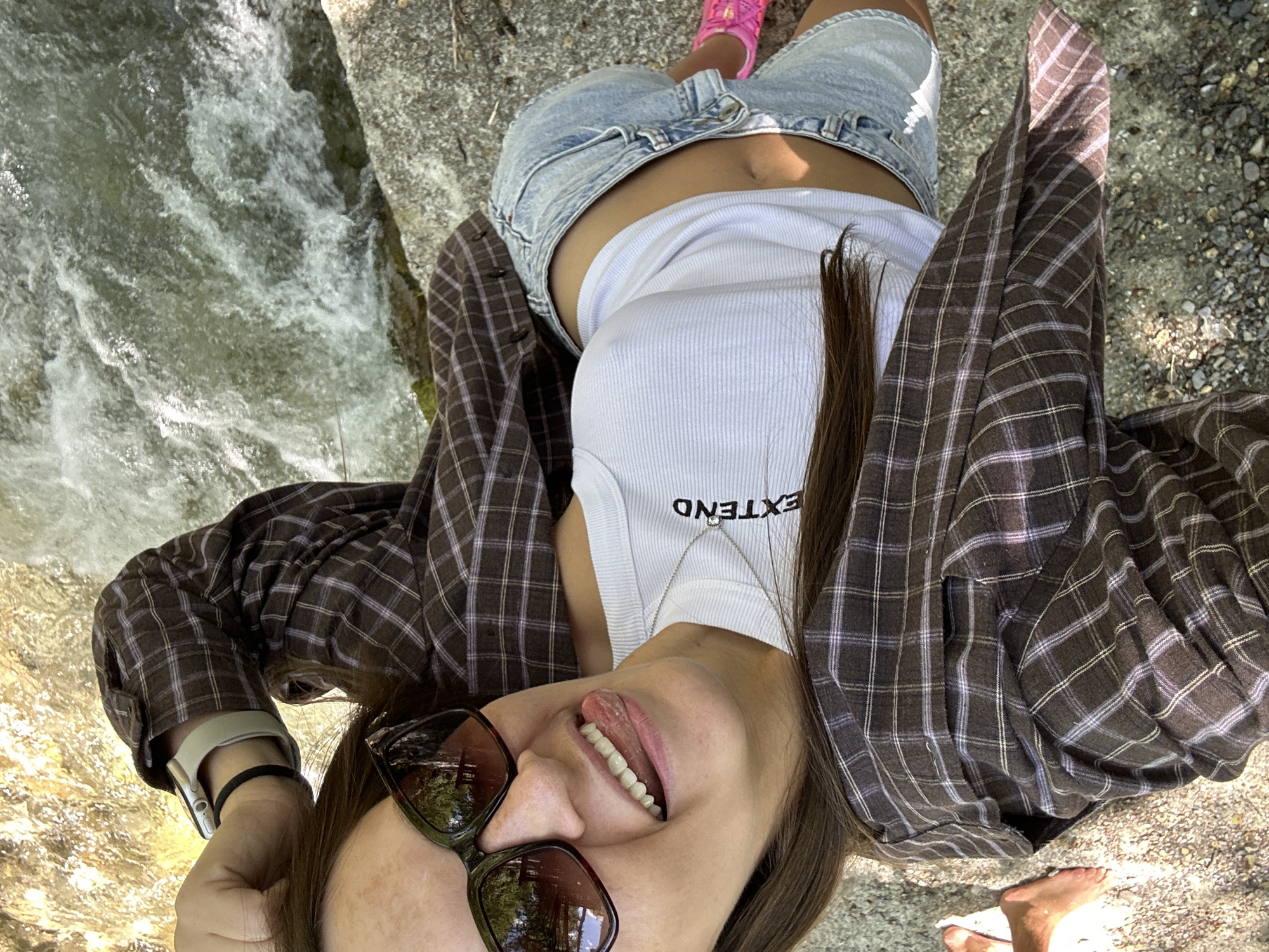 A young woman with long, brown hair is lying on her back next to a body of water, possibly a stream or river. She is wearing a white tank top with the word "EXTEND" printed on it, denim shorts, a brown and purple plaid button-up shirt worn open, and pink shoes. She is also wearing sunglasses and a watch on her left wrist. Her tongue is sticking out and her mouth is open. The water appears to be moving and shallow, with rocks and pebbles visible in and around it. The ground beneath her is rocky and sandy. The overall impression is a casual, outdoor scene. The location is not specified in the image or description.