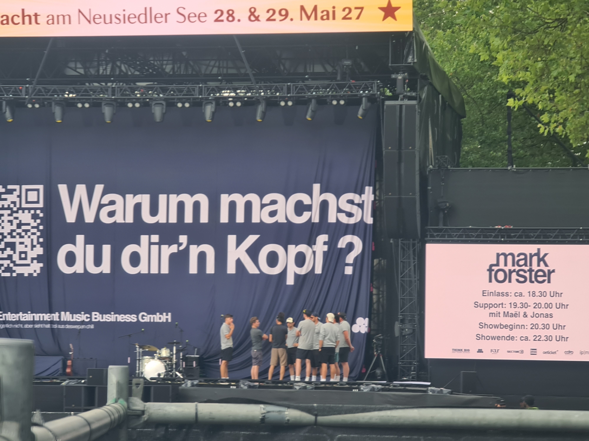 The image captures a daytime outdoor event stage setup, likely a concert or festival, with a large blue banner as the backdrop. The banner displays large white German text: "Warum machst du dir'n Kopf?". To the left of the banner, a QR code is visible. Below the main text, smaller white text reads "Entertainment Music Business GmbH" followed by some illegible German text.
In the foreground, on the stage, a group of men in casual attire, mostly t-shirts and shorts, are gathered. They appear to be staff or artists, some wearing baseball caps. Some are talking to each other, while others are standing and observing. A drum kit and a violin are visible on the stage, suggesting a musical performance is imminent or has recently concluded.
To the right of the main banner, a large screen displays event information for "mark forster". It lists "Einlass: ca. 18.30 Uhr", "Support: 19.30- 20.00 Uhr mit Maël & Jonas", "Showbeginn: 20.30 Uhr", and "Showende: ca. 22.30 Uhr". Below this, several logos are displayed.
The overall environment suggests a public gathering with a focus on live entertainment. The presence of trees in the background indicates an outdoor venue. Based on the text on the screen, the event is likely a concert by the artist Mark Forster. While the image itself does not explicitly state the location, searching for "Mark Forster Klagenfurt" and considering the text "Nacht am Neusiedler See" on the top banner, the event is likely a music festival or concert held near the Neusiedler See, possibly in the general vicinity or in connection with an event in Klagenfurt, Austria, although the specific venue cannot be definitively identified from the image alone.