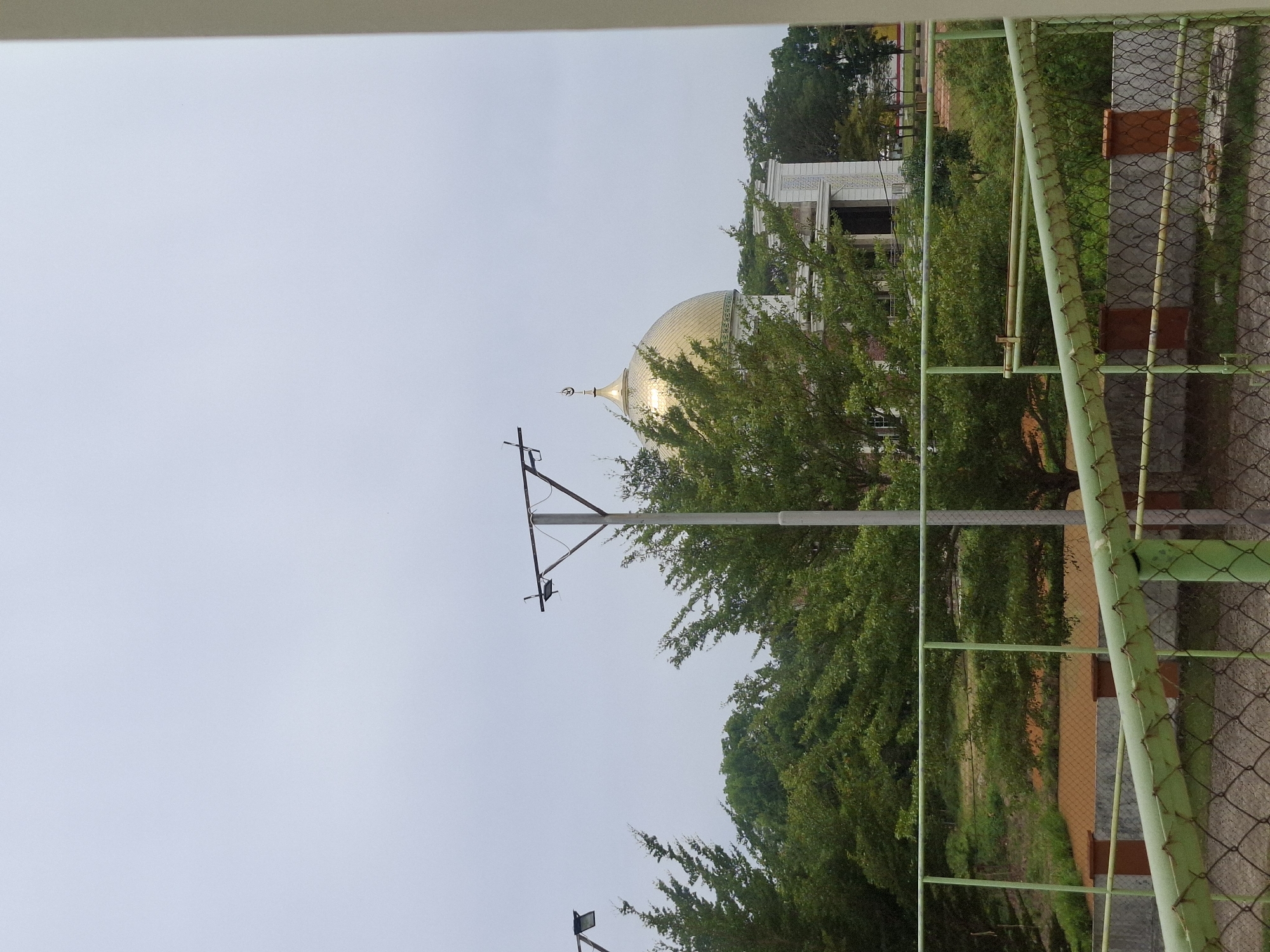 The image shows a mosque with a golden dome, likely located in Meureudu, Indonesia, given the architectural style and common features of mosques in the region. The mosque is surrounded by lush green trees, and in the foreground, there is a green metal fence and a chain-link fence. The sky is overcast, suggesting a cloudy day. In the distance, there are streetlights, and the overall setting appears to be an outdoor area, possibly a park or public space adjacent to the mosque. The image captures a serene and tranquil scene, with the mosque being the central focal point.