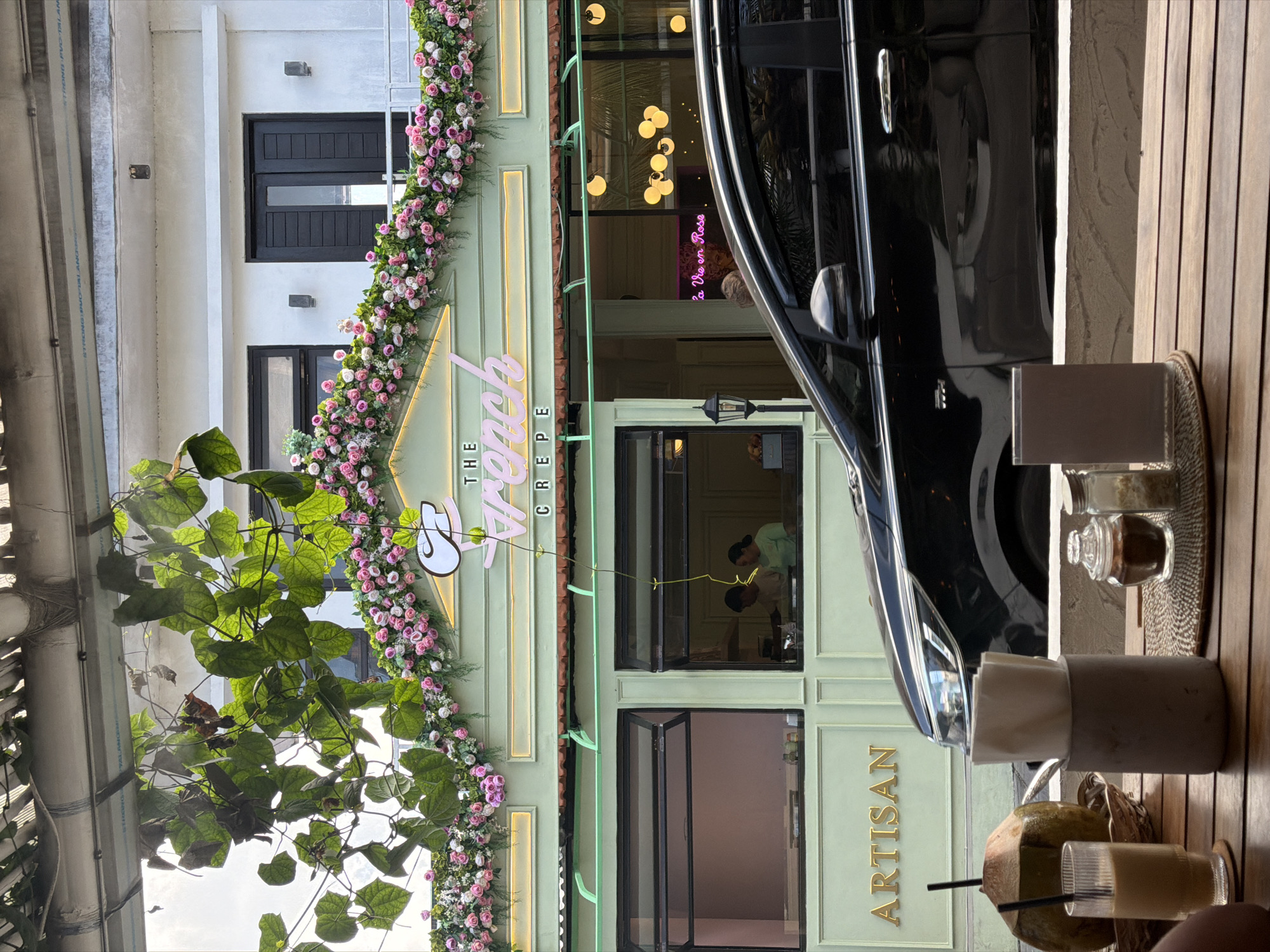 This image captures an outdoor seating area in front of "The French Crepe," a restaurant located in North Kuta, Indonesia. The facade of the restaurant is painted a light mint green and is adorned with a vibrant floral garland of pink and white roses. The name of the establishment is illuminated in neon pink. Below the garland, large windows reveal the interior, where two individuals, likely staff, are visible. One window has a pink-tinted interior, while the other shows a more neutral setting. To the left of the windows, the word "ARTISAN" is displayed in gold lettering.
In the foreground, a wooden table is set with various items. A halved coconut with straws sits in a woven basket. Next to it, a tall glass contains a light-colored beverage. A concrete holder with napkins is also present, alongside small glass jars containing what appear to be sugar and a dark condiment. A small, dark brown card rests on a woven placemat.
A black car is parked partially in front of the restaurant, obscuring some of the lower facade and the sidewalk. The overall scene suggests a relaxed, cafe-like atmosphere, possibly during daytime hours, given the natural light.