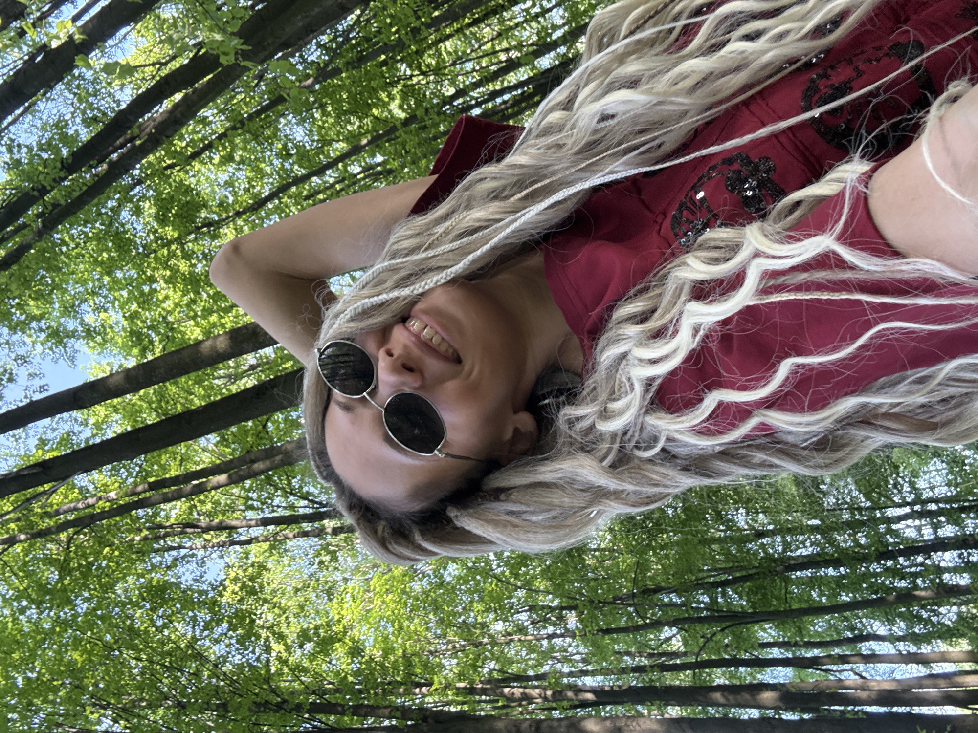 A woman wearing sunglasses and a maroon dress with decorative embellishments is photographed from a low angle, appearing to be lying down looking up. Her head is tilted, and she is smiling. Her hair is long and blonde with interspersed white braids. The background consists of a dense forest canopy with tree trunks visible. The location is not discernible as Rusanovo, Russia, from the image.
