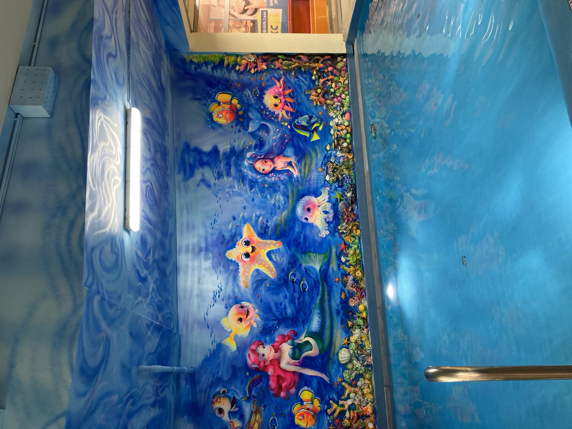 The image depicts an enclosed space with an underwater mural. On the left wall, a blue and white design mimics water currents, with a rectangular light fixture mounted. The central wall features a colorful mural of cartoon sea creatures, including fish, a starfish, a mermaid, and coral. The right wall is painted a solid blue, resembling water, and a metal handrail is visible. This underwater-themed decoration is located in the city of Kazan, Russia.