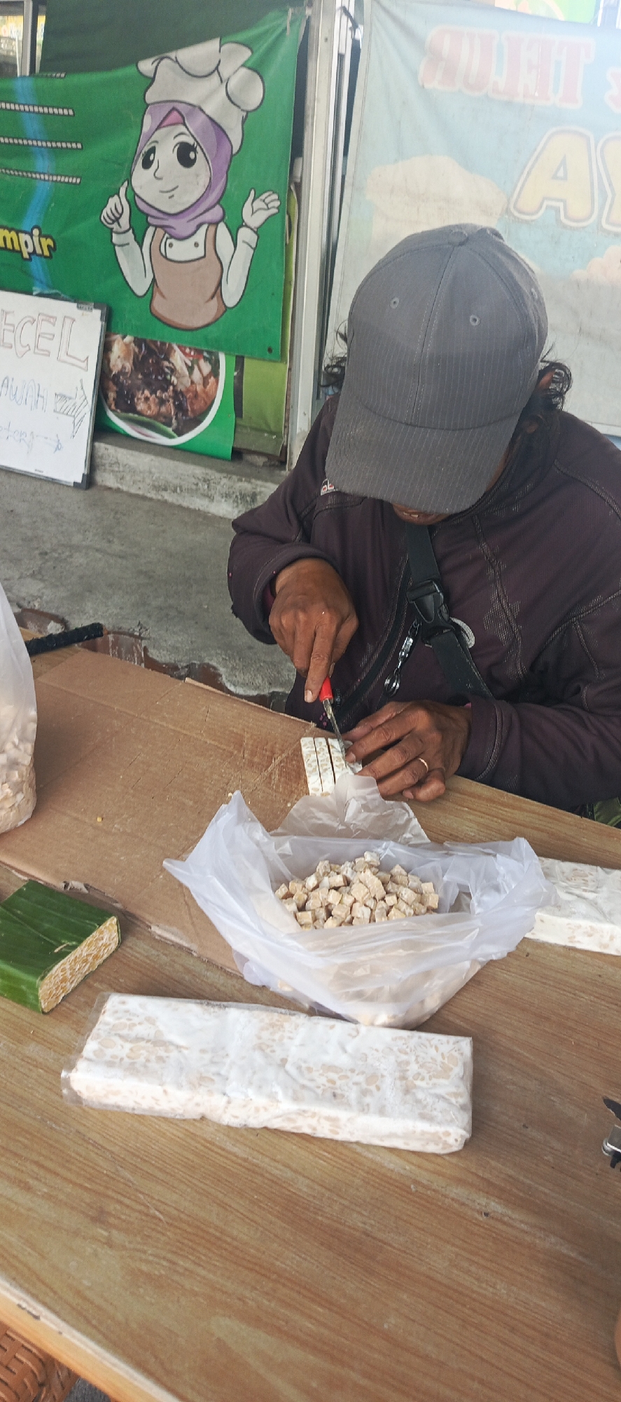 In Sukaresmi, Indonesia, a person wearing a gray baseball cap and a dark jacket is seated at a wooden table, actively engaged in slicing tempeh with a small knife. The tempeh is being prepared in small cubes. To the left of the person, a large piece of tempeh is wrapped in green banana leaves, and next to it, a white plastic bag holds a pile of diced tempeh. Another large block of tempeh, wrapped in plastic, sits further to the left on the table. In the background, a green banner with cartoon characters and text, possibly an advertisement, is visible, along with a white sign and another banner with light blue and white lettering. The overall environment appears to be an outdoor market or food stall.