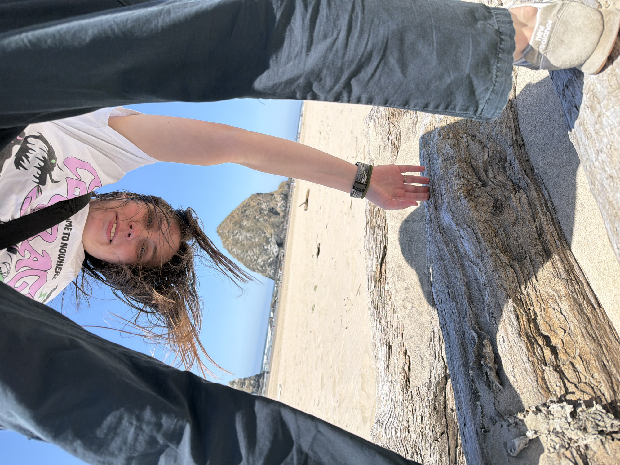 In Cannon Beach, Oregon, United States of America, a person is captured in an unusual, upside-down perspective on a sandy beach. The subject's head is positioned at the top of the frame, their long brown hair flowing around their face, which is looking downwards towards the camera. They are wearing a white t-shirt with a graphic print. Their right arm is extended downwards, with their hand resting on a large piece of driftwood in the foreground. A watch is visible on their wrist, and a ring on one of their fingers. The person's dark-colored pants frame the lower part of the image.
The background reveals the vast expanse of Cannon Beach, with its characteristic sandy shores and the iconic Haystack Rock jutting out of the ocean in the distance. The sky is a clear, bright blue, and the sun casts shadows on the sand and driftwood, indicating a sunny day. The overall impression is one of a playful and unique moment captured at this well-known coastal landmark.