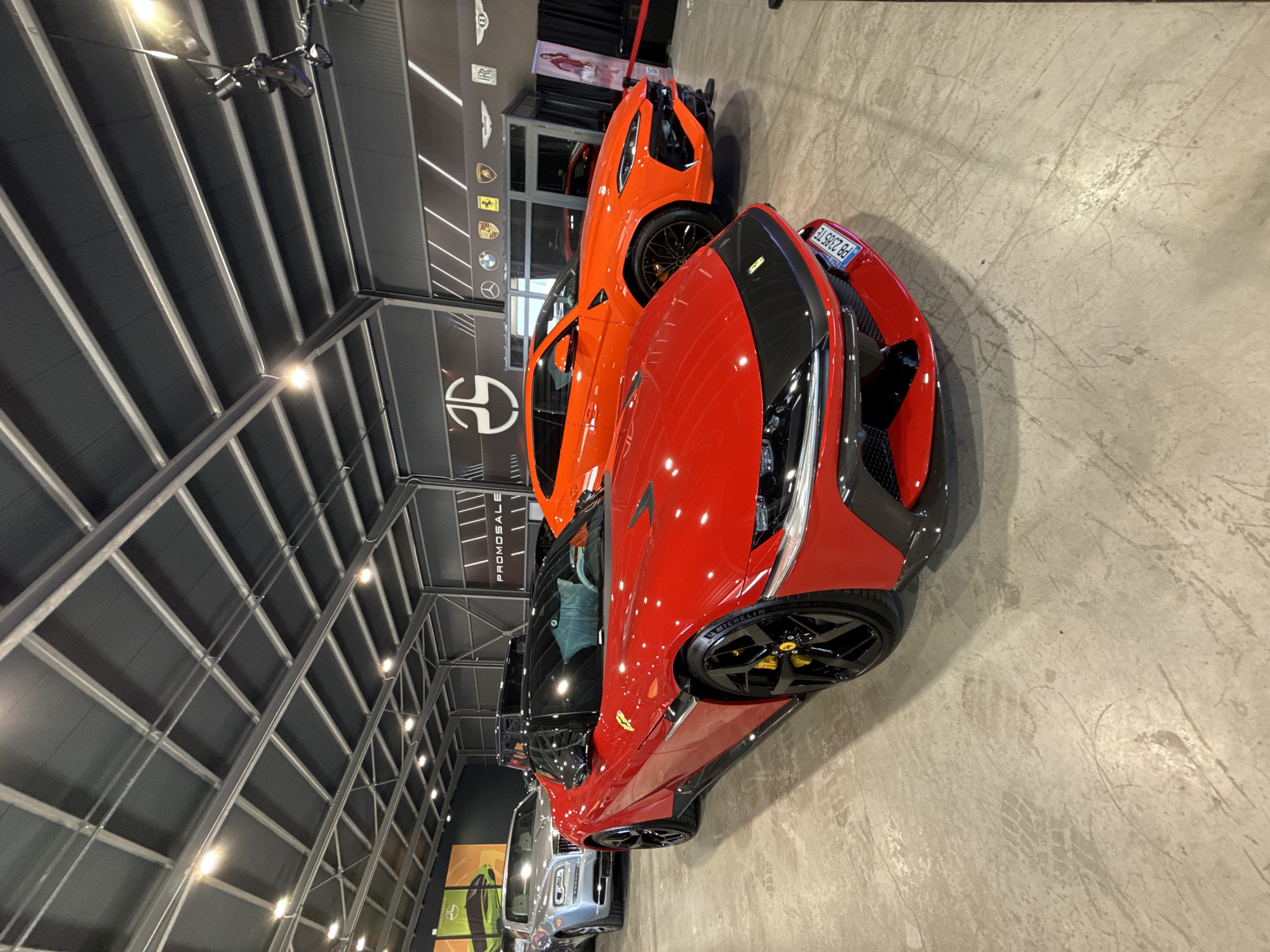 The image shows the interior of a car dealership. Multiple luxury vehicles are visible, including a red Ferrari with a black stripe and an orange Lamborghini. The Ferrari has a license plate that reads "PB 2305 TE." There is signage on the wall with a logo that appears to be of the dealership along with automotive company logos. The background reveals a metal-framed ceiling structure with lighting. The cars appear to be for sale. No people are visible. This dealership is located in the city of Plovdiv, Bulgaria.