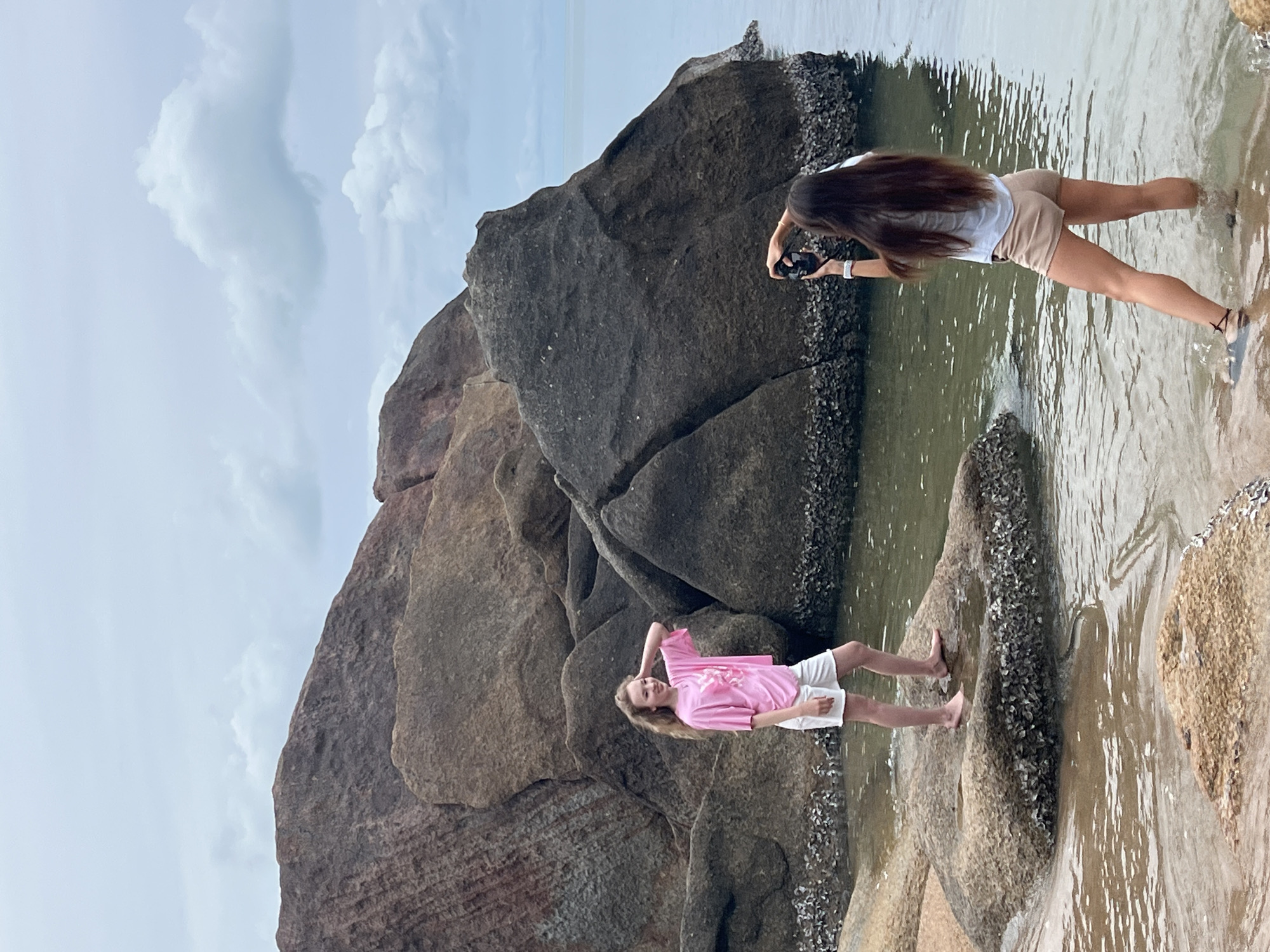 In Baan Chaweng Noi, Thailand, two young females are present on a rocky shoreline. The female on the left, with long blonde hair, is wearing a pink t-shirt and white shorts. She is standing on a rock, looking towards the right, with her right hand touching her head. The female on the right, with long dark hair, is wearing a light blue t-shirt and tan shorts. She is facing away from the camera, holding a black camera in her hands, and appears to be taking a photo of the other female. They are standing in shallow, clear water amongst various large, dark-colored rocks that are partially submerged. The sky above is cloudy and overcast, with the ocean visible in the background. The overall scene depicts a casual moment of photography at the beach.