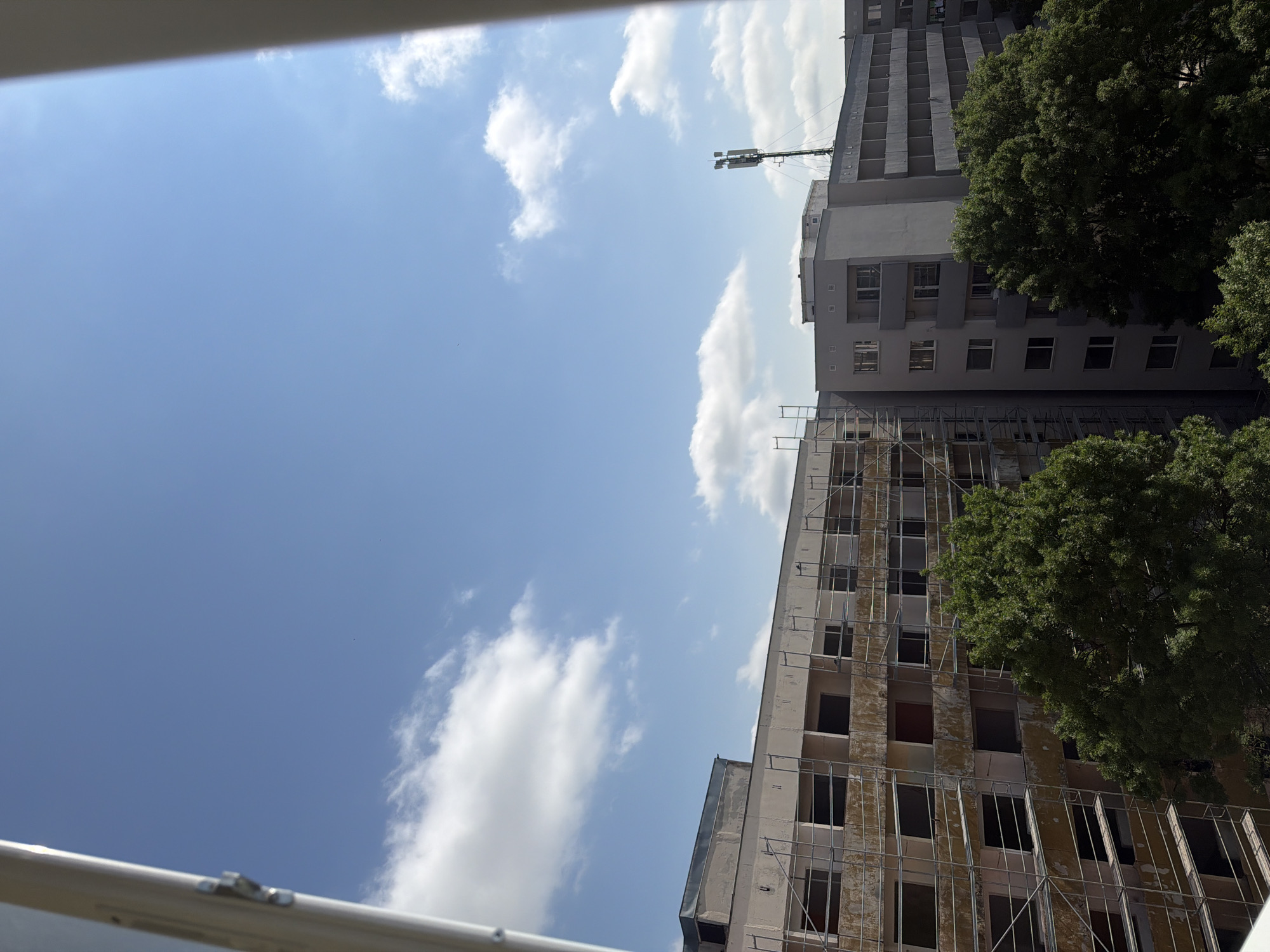 This image depicts an urban landscape in Sofia, Bulgaria, showcasing a mix of buildings, natural elements, and a bright sky. The perspective, taken from what appears to be a window or balcony, is oriented vertically but the image is presented rotated 90 degrees clockwise.
In the upper left quadrant of the properly oriented image, a clear, vibrant blue sky dominates, interspersed with scattered, fluffy white clouds. The sun casts bright light, indicating daytime, likely late morning or early afternoon.
Towards the right side of the frame, two multi-story buildings are visible. The more prominent building in the foreground, stretching upwards from the lower right, is light-colored, possibly beige or pale grey. Its facade is covered by an extensive scaffolding or metal framework, suggesting ongoing renovation or construction. Many of its window openings are dark and empty, revealing unfinished interiors. Behind this structure, a taller, darker grey building stands, appearing more complete with uniform rows of rectangular windows. A telecommunication antenna or cell tower is mounted on the roof of this rear building, extending into the clear sky.
Lush, dense green foliage from several trees partially obscures the right side of both buildings, adding a natural contrast to the concrete structures. A white or light-colored horizontal bar, possibly part of a window frame or balcony railing, is visible along the very bottom edge of the image. No people are visible, and there is no discernible text within the scene. The overall impression is a snapshot of urban development or renewal under a bright, clear sky.