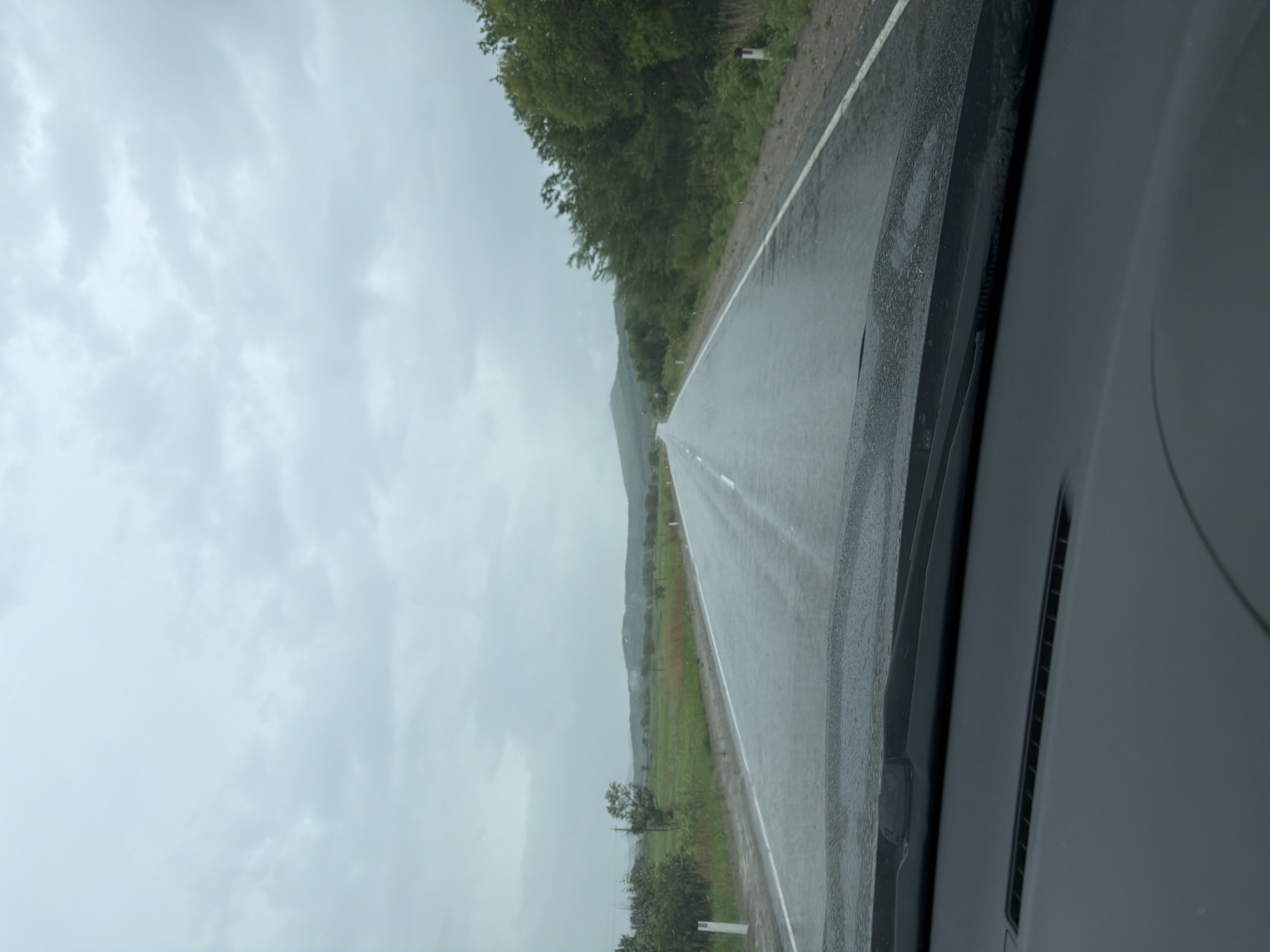 The image, captured from inside a vehicle, depicts a wet asphalt road extending into the distance under an overcast sky. The perspective is looking forward from the driver's or passenger's seat. The road surface gleams with moisture, reflecting the light from the sky, indicative of recent or ongoing rainfall. Faint white road markings are visible on the damp pavement.
To the left of the road, a dense row of green trees lines the route, beyond which lie expansive green fields. In the hazy distance, a large, undulating hill or mountain range emerges, its contours softened by the cloudy conditions. A small white roadside marker with a red top is subtly visible amidst the greenery on the left.
Sections of the vehicle's dark gray dashboard and windshield frame occupy the lower edges of the frame. The sky is entirely covered by a blanket of light grey clouds, creating a uniformly overcast atmosphere. The scene conveys a sense of travel through a rural or natural landscape during damp, cloudy weather. There are no visible people or discernible text. The location context for this scene is Бабстово, Russia.