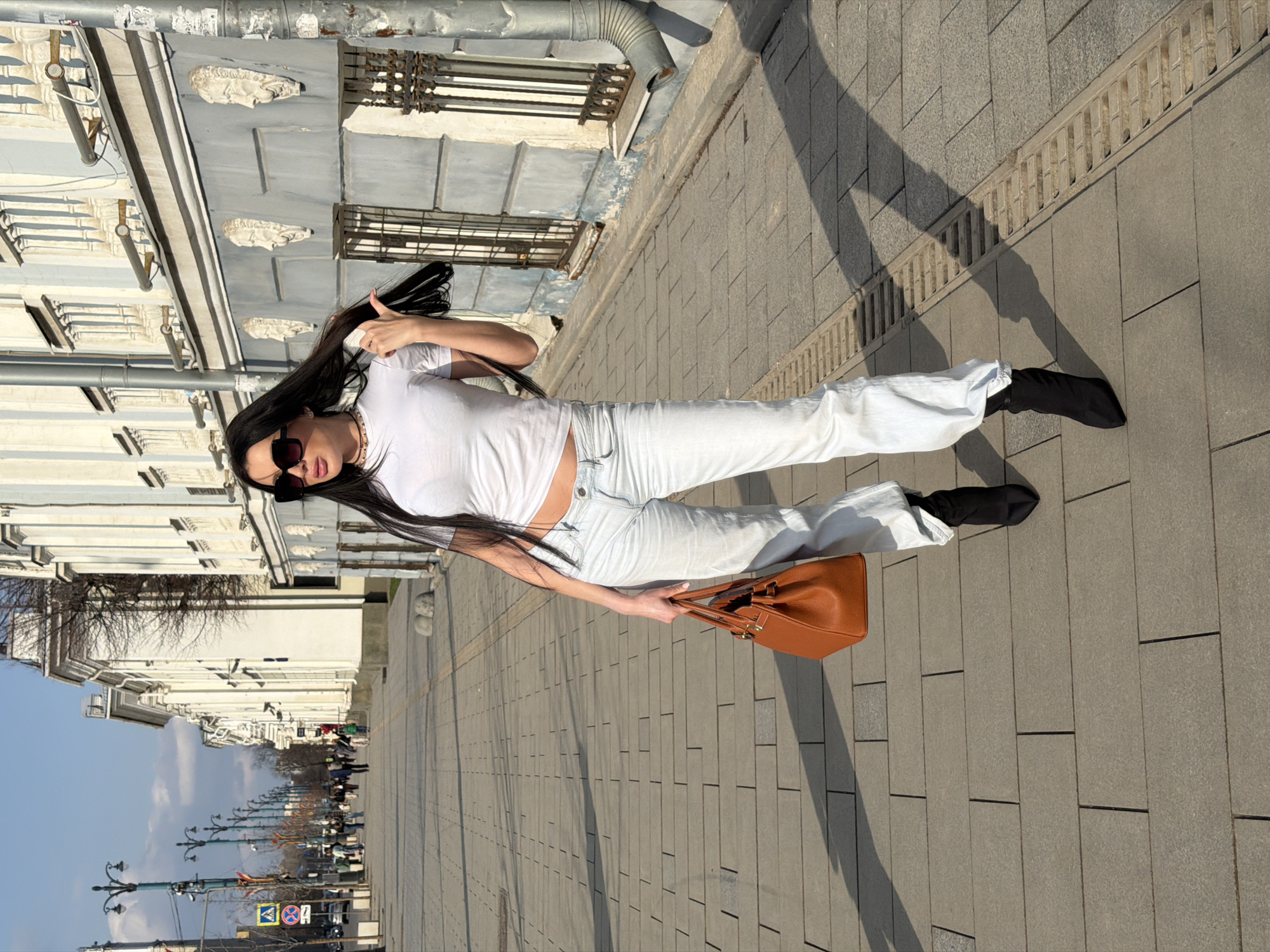 A young woman with long black hair, wearing sunglasses, a white t-shirt, light blue jeans, and black boots, stands on a wide sidewalk in Voronezh, Russia. She is holding a brown handbag and giving a thumbs-up. The sidewalk is made of gray rectangular pavers. A building with multiple windows is on her left and the street is on her right. Street lights and pedestrians can be seen further down the sidewalk in the background. The sky is blue.