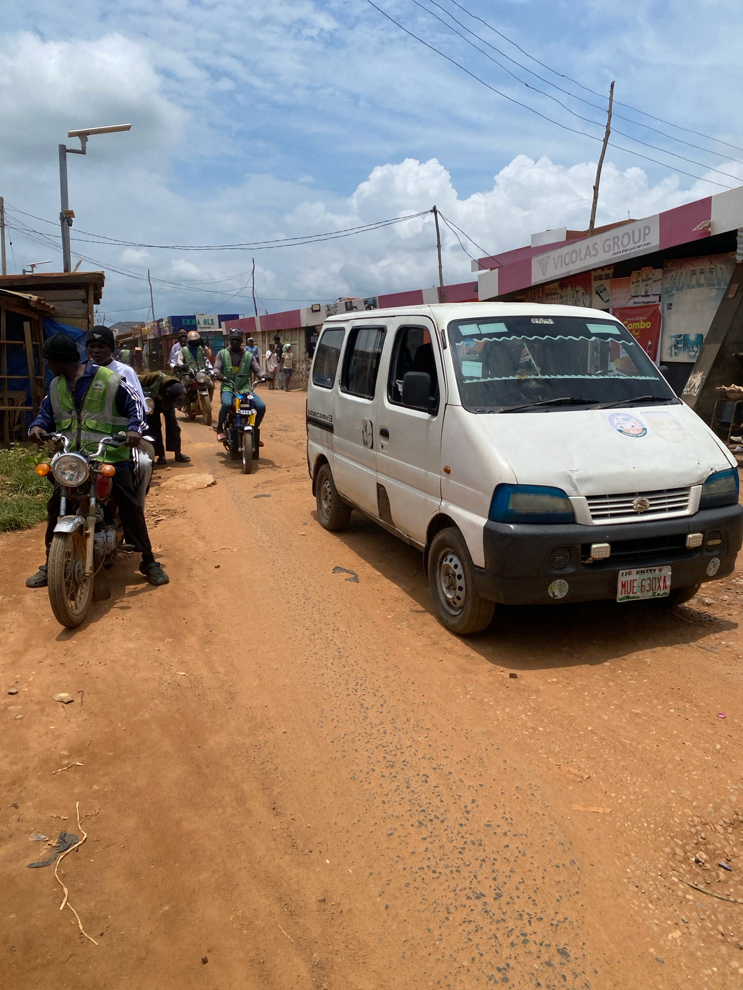 In Iworoko, Nigeria, a white Suzuki Carry van is parked on a dirt road. A man in a yellow vest rides a motorcycle in the foreground, with another man in a similar vest behind him on the same motorcycle. A third man on a motorcycle is visible in the background. Buildings line the side of the road, one with a sign for "VICOLAS GROUP." The sky is cloudy.