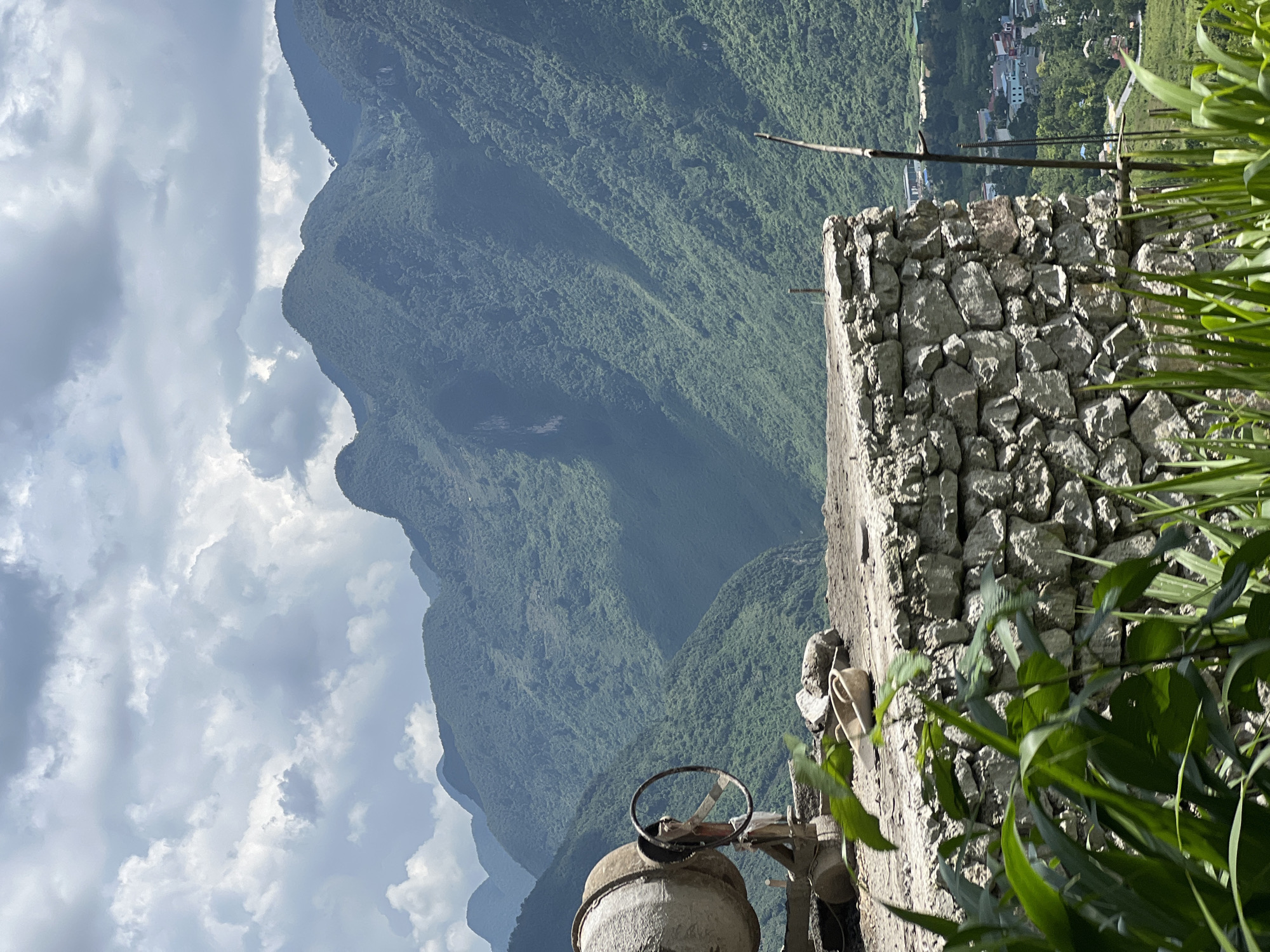 The image depicts a construction scene in Lùng Tám Commune, Vietnam, set against a backdrop of lush, verdant mountains under a cloudy sky. In the foreground, a concrete mixer is positioned on a constructed platform made of rough-hewn stones. The platform is topped with a layer of dirt or gravel, upon which a small trowel and some rocks are scattered. Lush green foliage, possibly corn stalks, partially obscures the lower portion of the scene. In the mid-ground, the stone wall of the platform is prominent, with a wooden stick or stake planted vertically next to it. Beyond the platform, the rolling, densely forested mountains dominate the landscape, their slopes carved by valleys and shadows. In the distance, a small village with scattered buildings is visible nestled amongst the trees at the foot of the mountains. The sky is filled with dramatic, heavy clouds, suggesting an overcast or potentially stormy day. There are no people visible in the image.