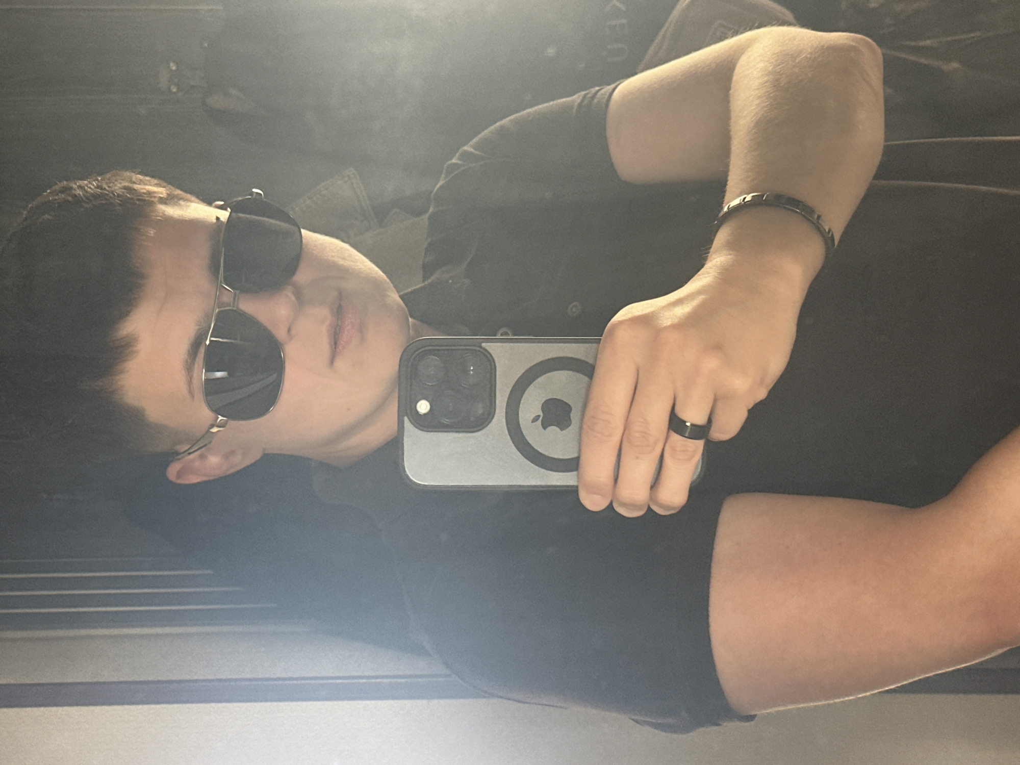 A person is taking a mirror selfie in what appears to be a dimly lit room. The person is wearing a black polo shirt, aviator sunglasses, and a black ring on their left ring finger. They are holding a smartphone in their left hand, which has a case with an Apple logo. A silver-toned bracelet is visible on their right wrist. The background is blurry, but suggests a closet or wardrobe area with some clothing items. There is a vertical line on the left, possibly a doorframe or wall edge. The overall lighting is low, with a strong light source to the left creating a glare. The image does not contain any identifiable landmarks or features that would indicate a specific location in Uzhhorod, Ukraine.
