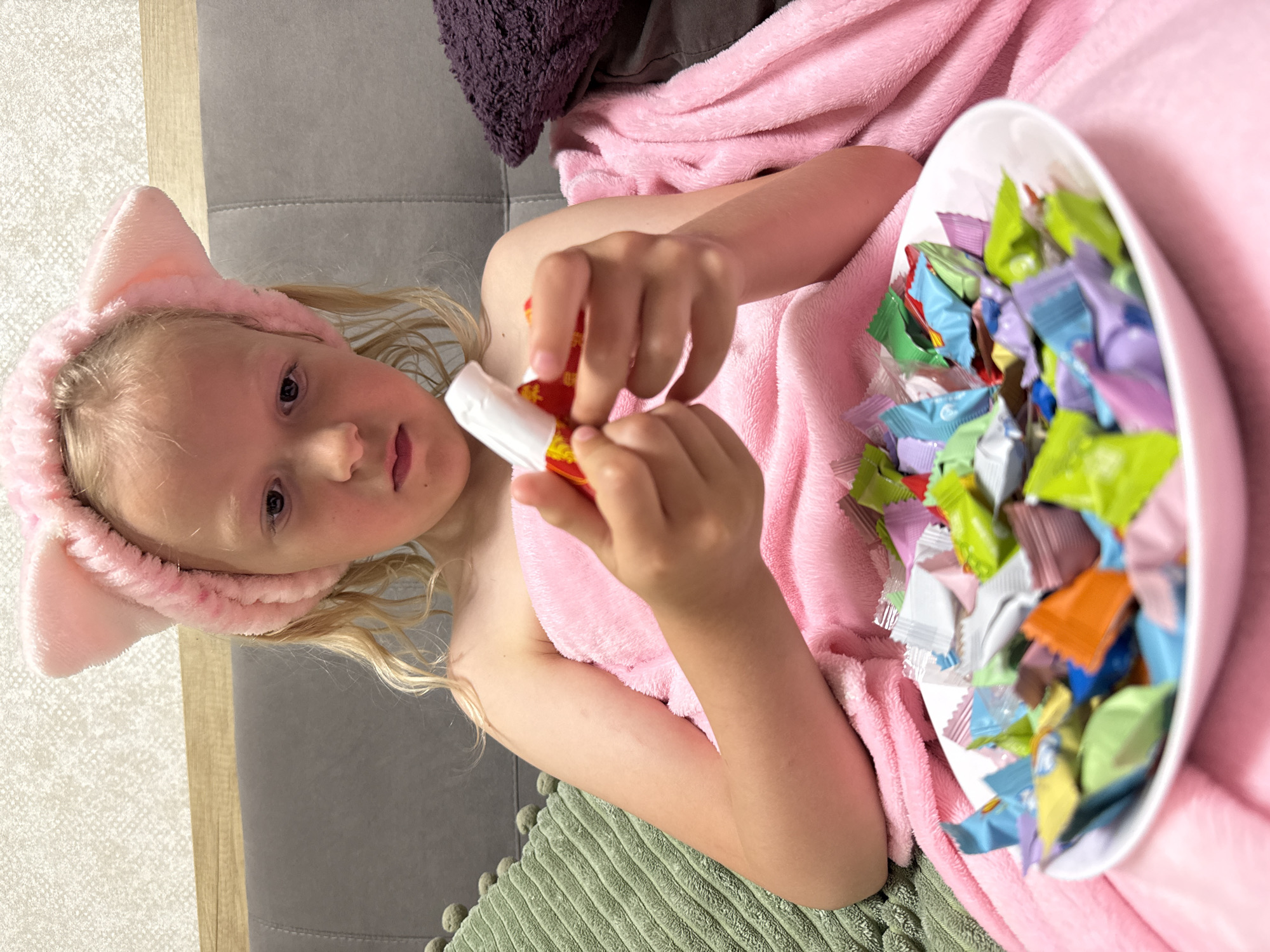 A young fair-skinned child, possibly female, lies on a grey couch. The child wears a pink headband with ears and is covered in a pink blanket. They are holding a red and white item in their hands. To the right of the child is a white bowl filled with numerous individually wrapped candies of various colors. Part of a green rug is visible under the child's arm. To the upper left of the child's head is a purple pillow. The location is Unknown, Unknown.