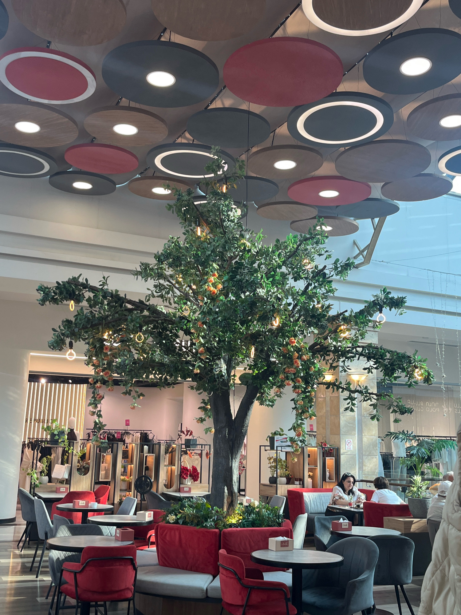 The image depicts the interior of a modern cafe, likely located in Yerevan, Armenia, given the context provided. The ceiling features a distinctive design of large, circular panels, some illuminated from within, in shades of grey, red, and wood tones, suspended at varying heights. A prominent, artificial tree with green foliage and decorative elements like small orange fruits and hanging lights is centrally positioned in the cafe.
The cafe's seating area is furnished with a mix of comfortable sofas and chairs in red and grey upholstery, arranged around round tables. Some tables have small boxes of tissues or other decorative items on them. In the background, there's a retail display area with mannequins and clothing racks, suggesting the cafe might be part of a larger establishment or a retail space with an integrated cafe.
Several patrons are visible in the cafe. Two women are seated at a table in the mid-ground, engaged in conversation. In the foreground, chairs and tables suggest a lively atmosphere, though the focus is on the overall ambiance and decor. The lighting appears to be a combination of ambient light from the ceiling fixtures and decorative lights, creating a warm and inviting environment. The overall scene conveys a sense of contemporary design and a relaxed social setting.