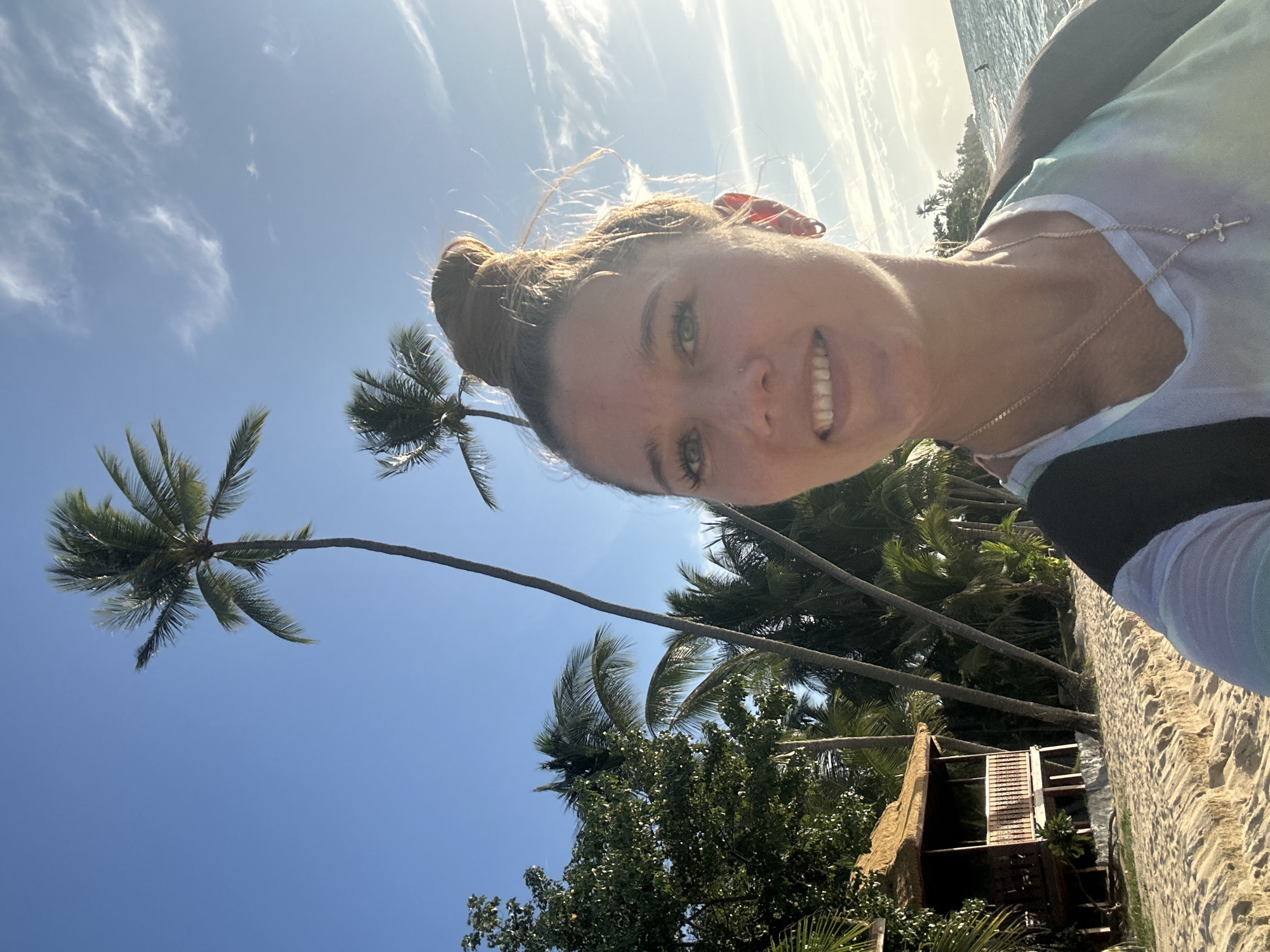 A woman with blonde hair in a bun smiles at the camera, taking a selfie on a sandy beach. She is wearing a white, blue, and purple tie-dye tank top and a silver cross necklace. Her right ear has two hoop earrings. Behind her, palm trees and a small hut with a thatched roof line the shore. The ocean is visible to the right, with a person swimming in the distance. The sky is a clear blue with some wispy clouds. The scene suggests a tropical location like Ko Samui District, Thailand, with its characteristic palm trees and beach huts.