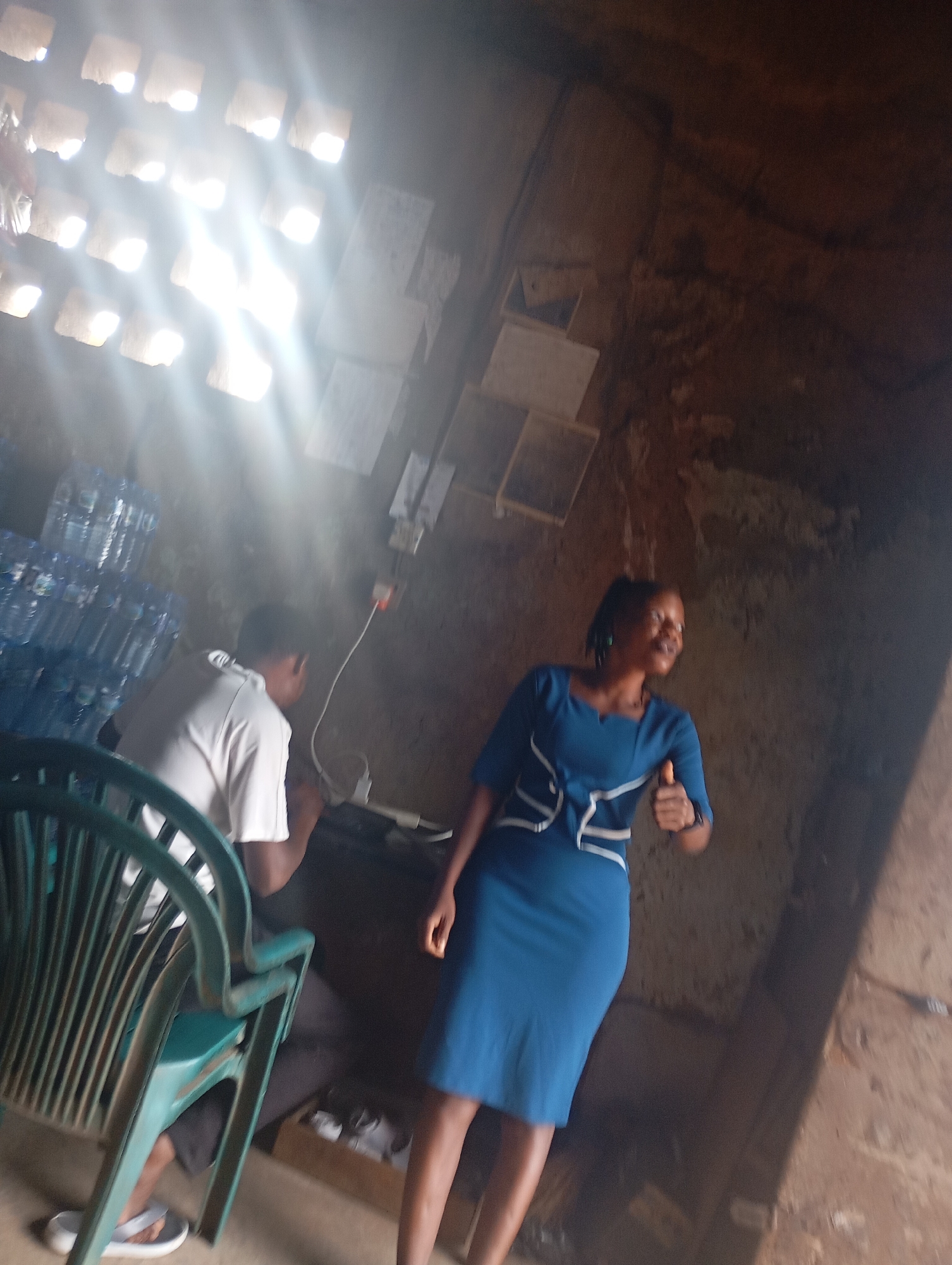 In an indoor setting, a woman in a blue dress stands and gives a thumbs up. To her left, a person in a white shirt is seated, facing away from the camera. In the background, stacks of plastic water bottles are visible. The room appears to be lit by natural light streaming through a window with multiple square openings. The walls are rough and textured, suggesting a rustic or unfinished environment.