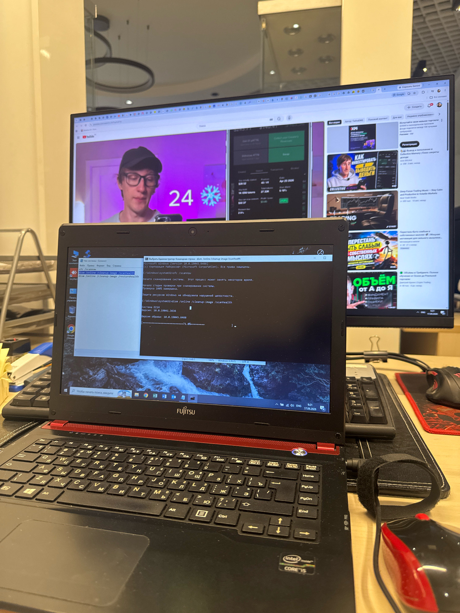 The image displays a desktop setup with two monitors and a laptop. The laptop, a Fujitsu model, is open and positioned in the foreground, showcasing its keyboard and screen which displays a command prompt window. The command prompt window contains text related to Windows system file checking and restoration. Behind the laptop, a larger monitor is visible, displaying a YouTube video with a person wearing glasses and a cap, with the number "24" and a snowflake icon. The right side of the larger monitor shows various web pages with Russian text, possibly related to finance or trading. A keyboard and a red and black computer mouse are situated on a desk mat to the right of the laptop. The overall environment appears to be an indoor office or home study space, with reflective surfaces suggesting it might be a well-lit area. There are no visible elements that definitively place the scene in Almaty, Kazakhstan; the context is limited to the digital content displayed and the objects present.